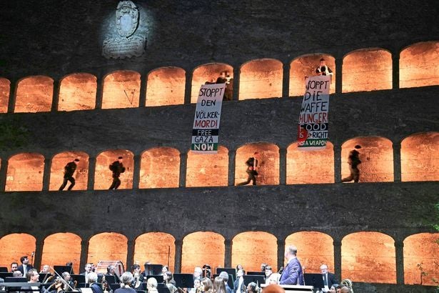 Protestplakate bei den Festspielen, Musiker spielen im Vordergrund, Personen im erleuchteten Gemäuer.