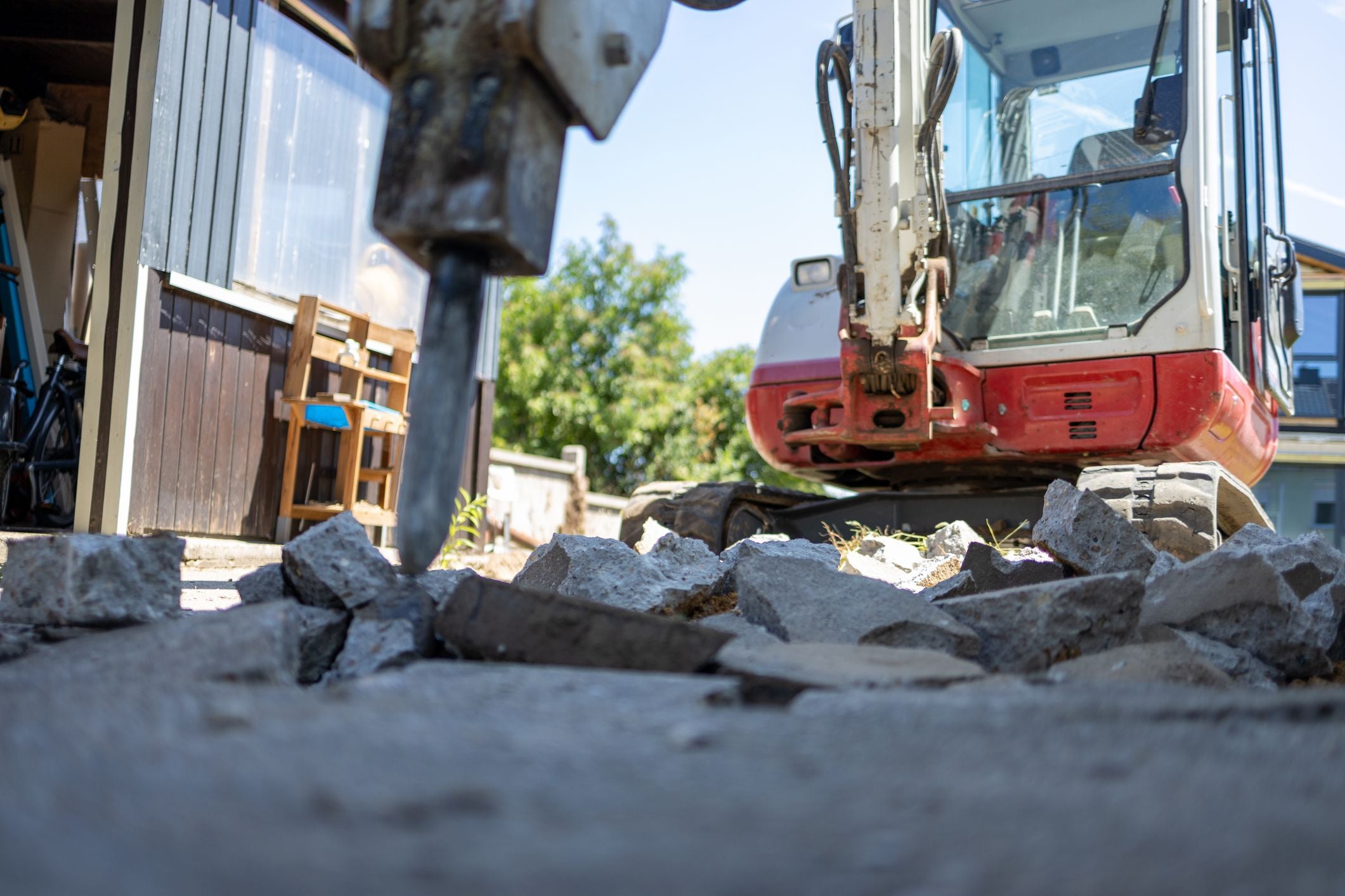 Ein Minibagger mit Hydraulikhammer zertrümmert Betonplatten vor einem Wohnhaus.
