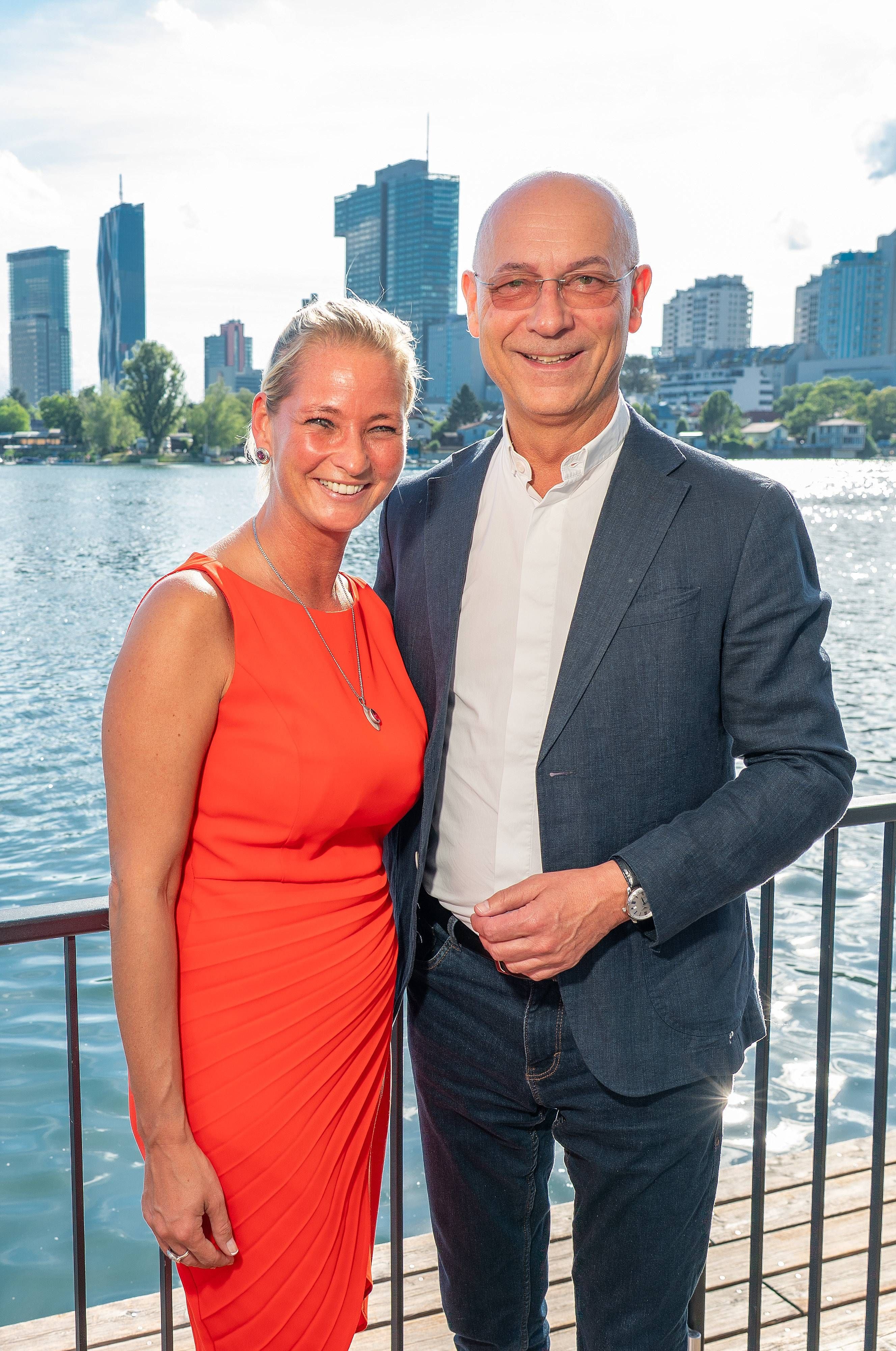 Ein Mann und eine Frau lächeln am Ufer der Alten Donau vor moderner Skyline in Wien.
