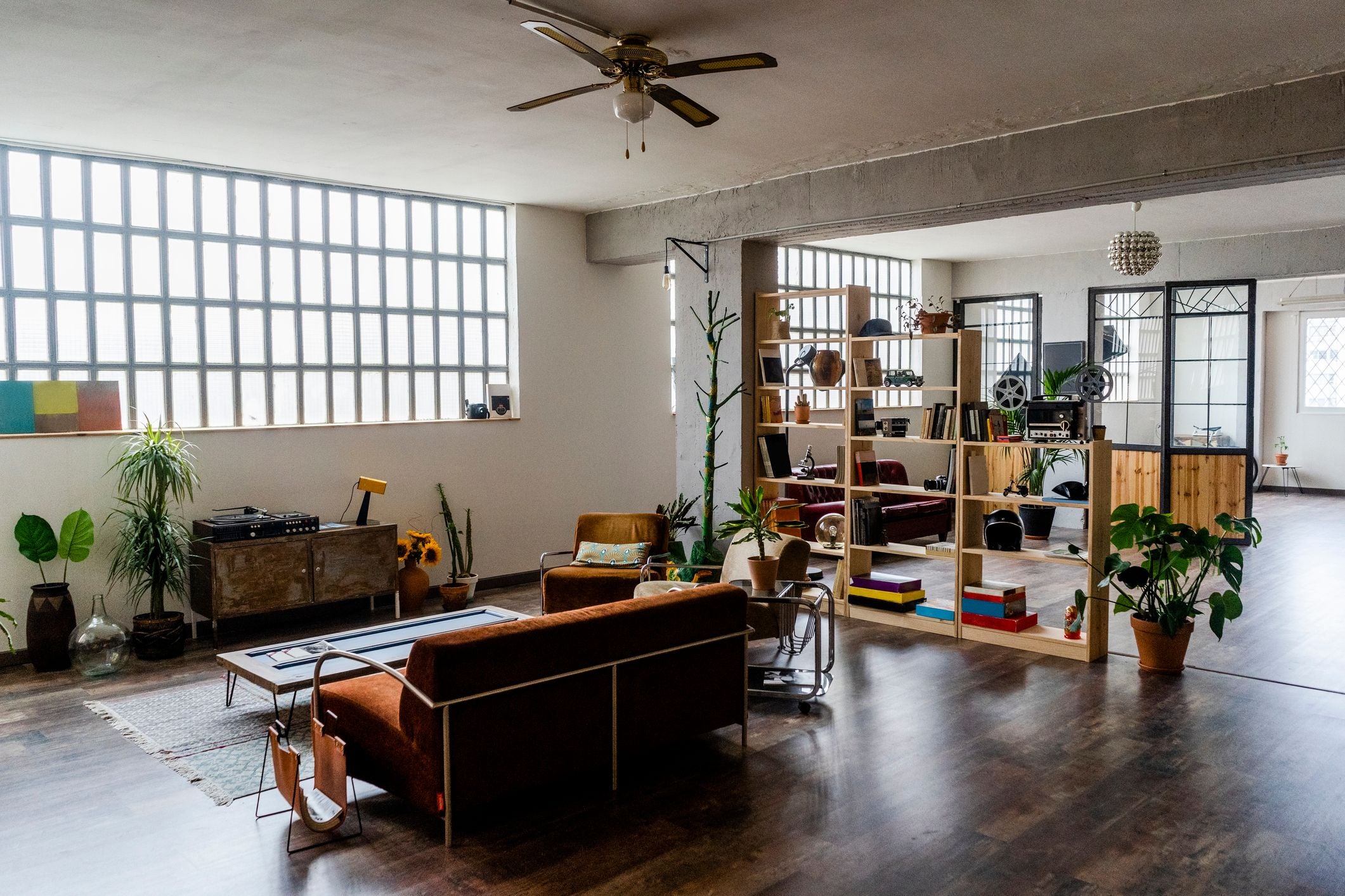 Modernes Loft-Wohnzimmer mit großen Fenstern, Pflanzen, Regalen und gemütlichen Sitzmöbeln.