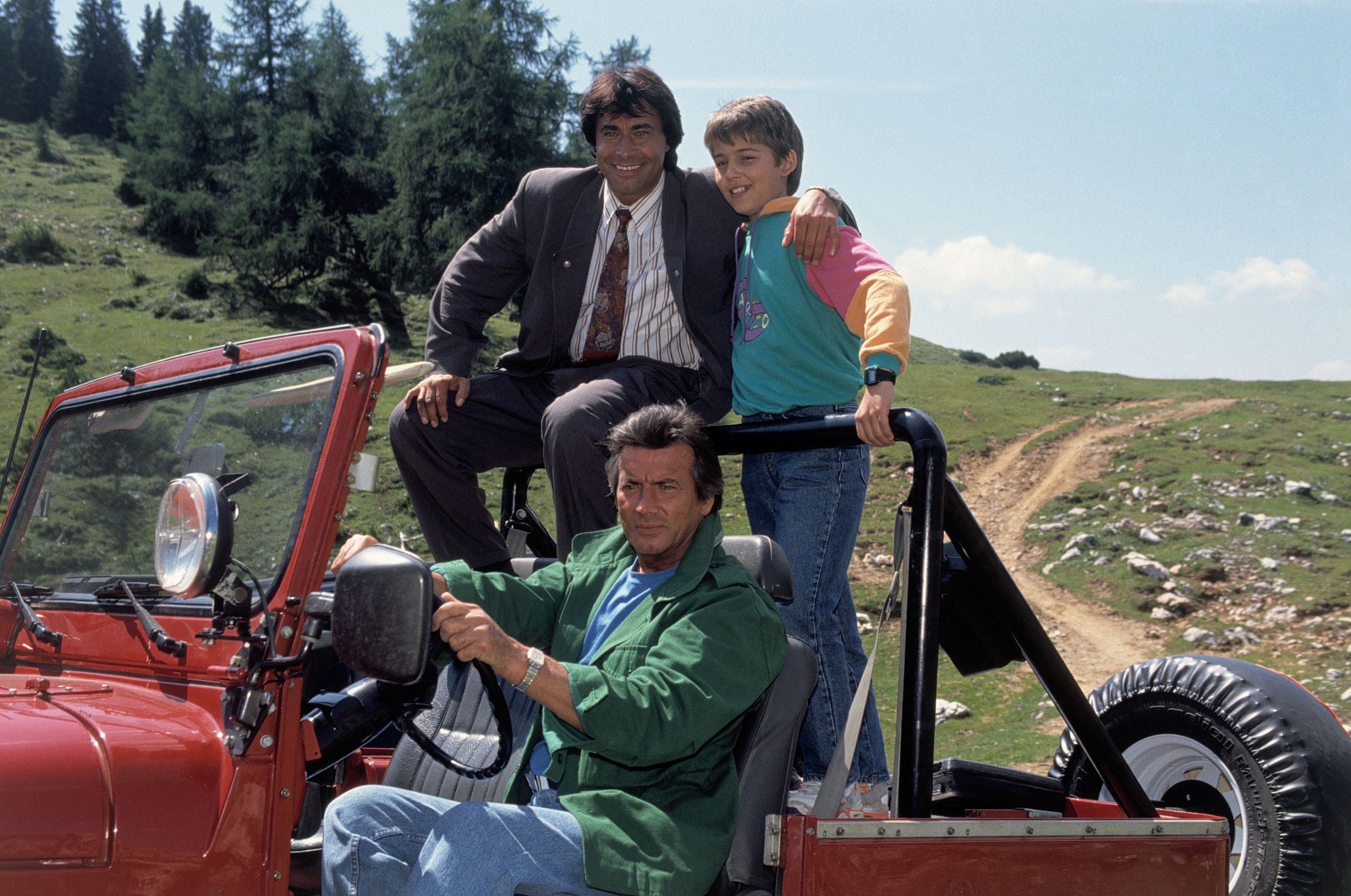 Drei Menschen auf einem roten Jeep vor einer grünen Hügellandschaft und Bäumen.