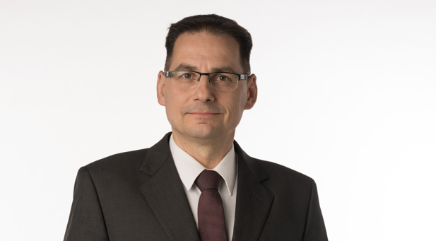 Mann mit Brille im dunklen Anzug und Krawatte vor hellem Hintergrund.