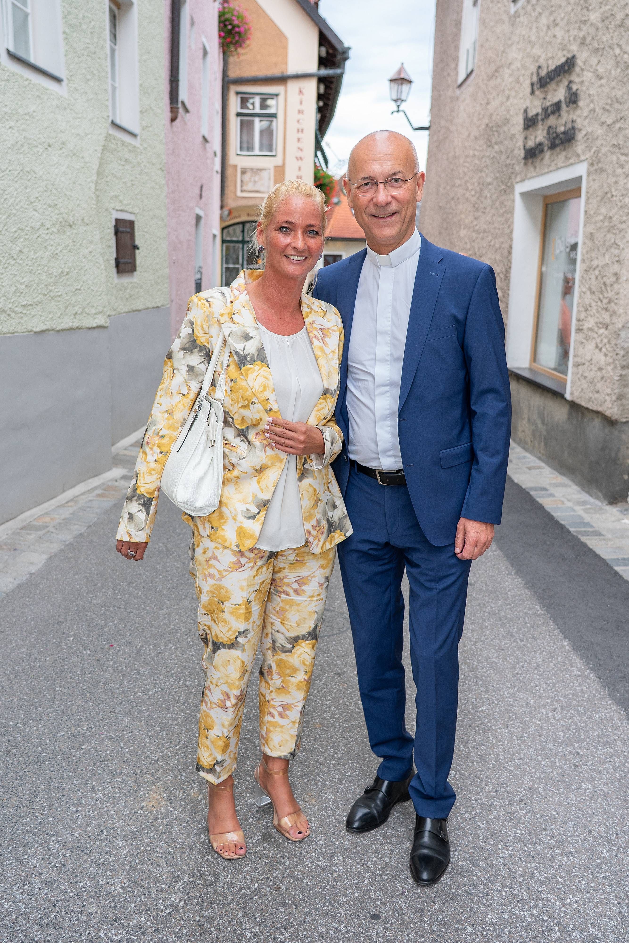 Ein lächelndes Paar steht elegant gekleidet auf einer Kopfsteinpflasterstraße.