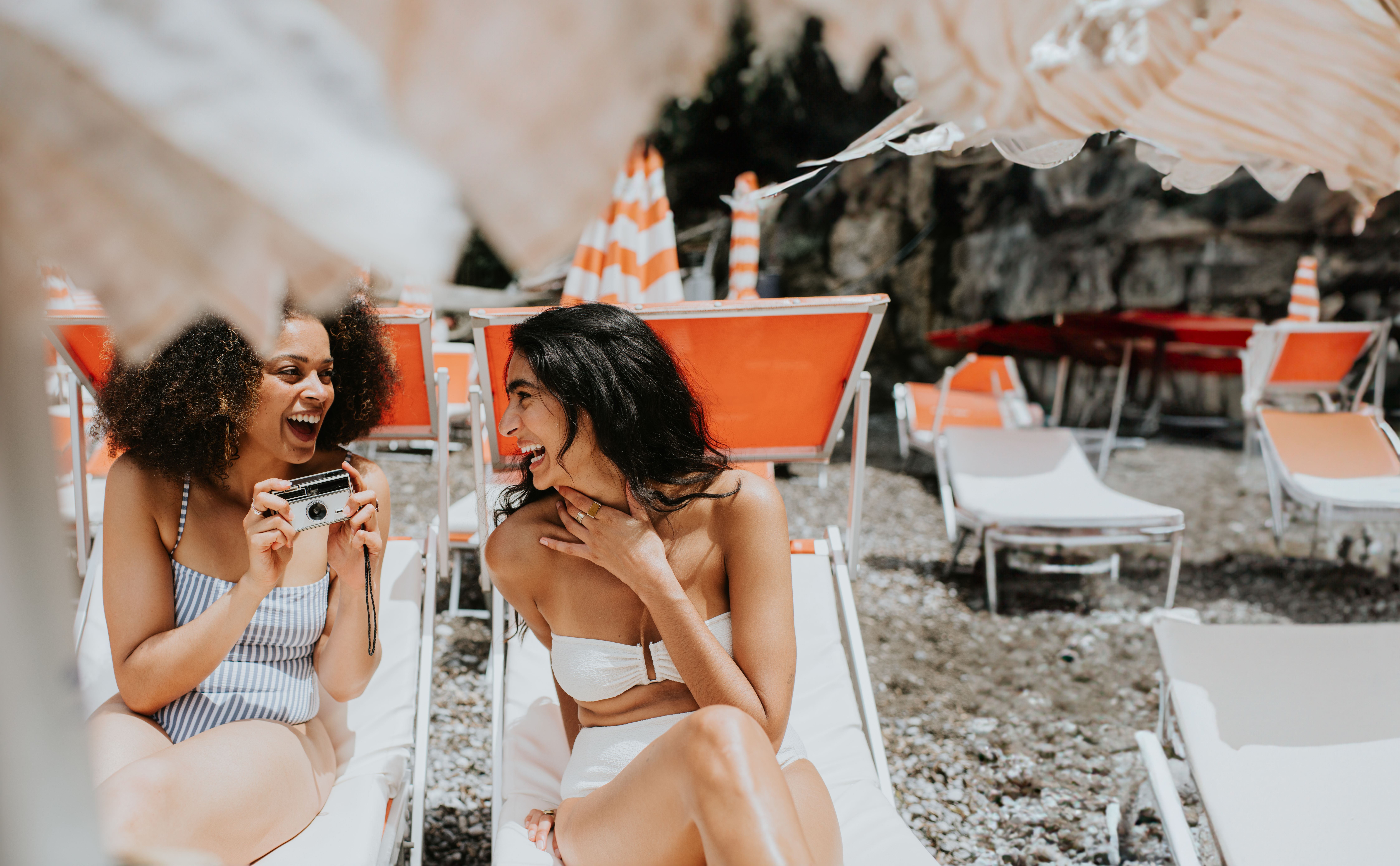 Zwei junge Frauen lachen gemeinsam auf Sonnenliegen in einem Ferienresort, eine hält eine Kamera.