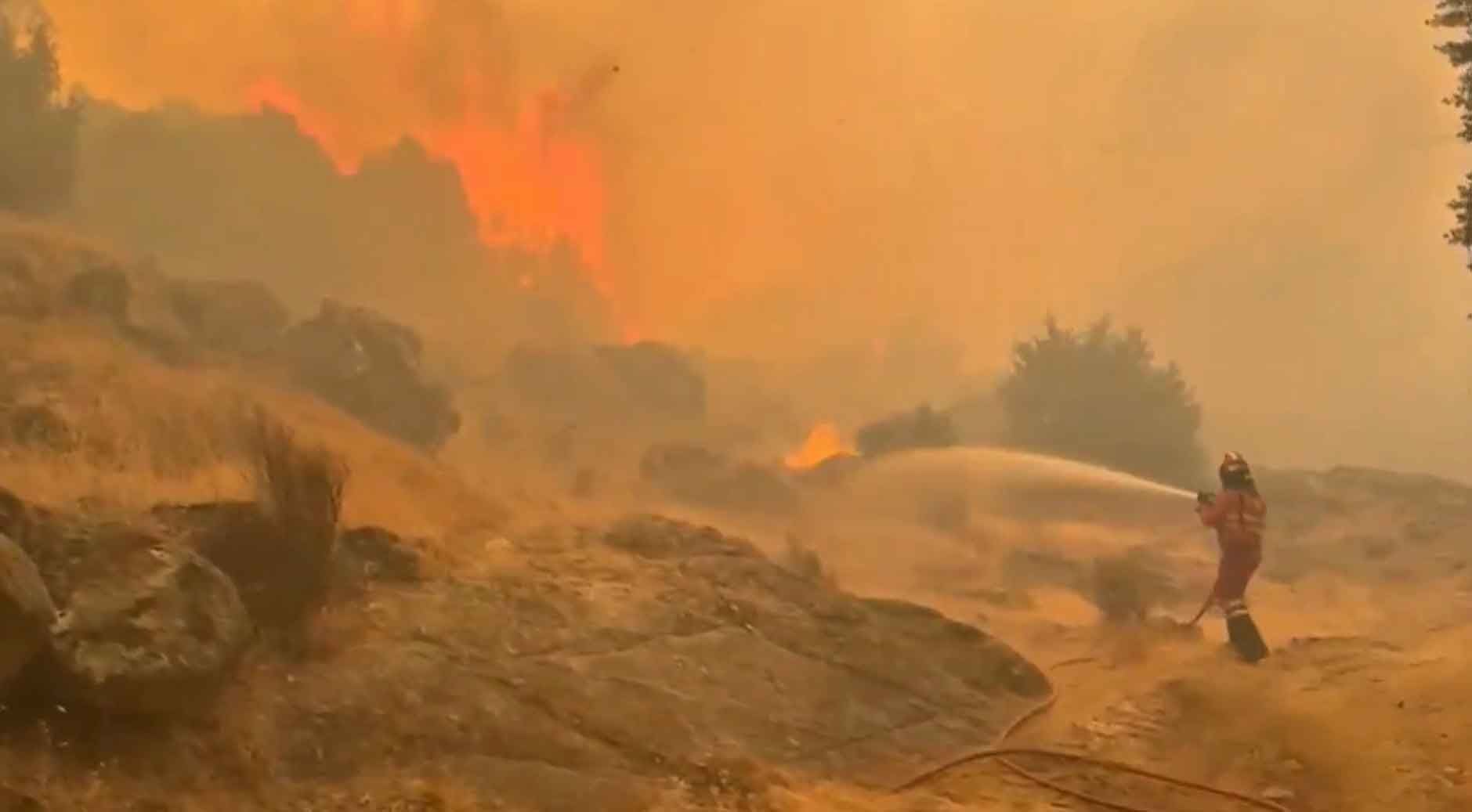 Feuerwehrmann bekämpft einen Waldbrand in Spanien mit einem Wasserschlauch, dichte Rauchentwicklung.