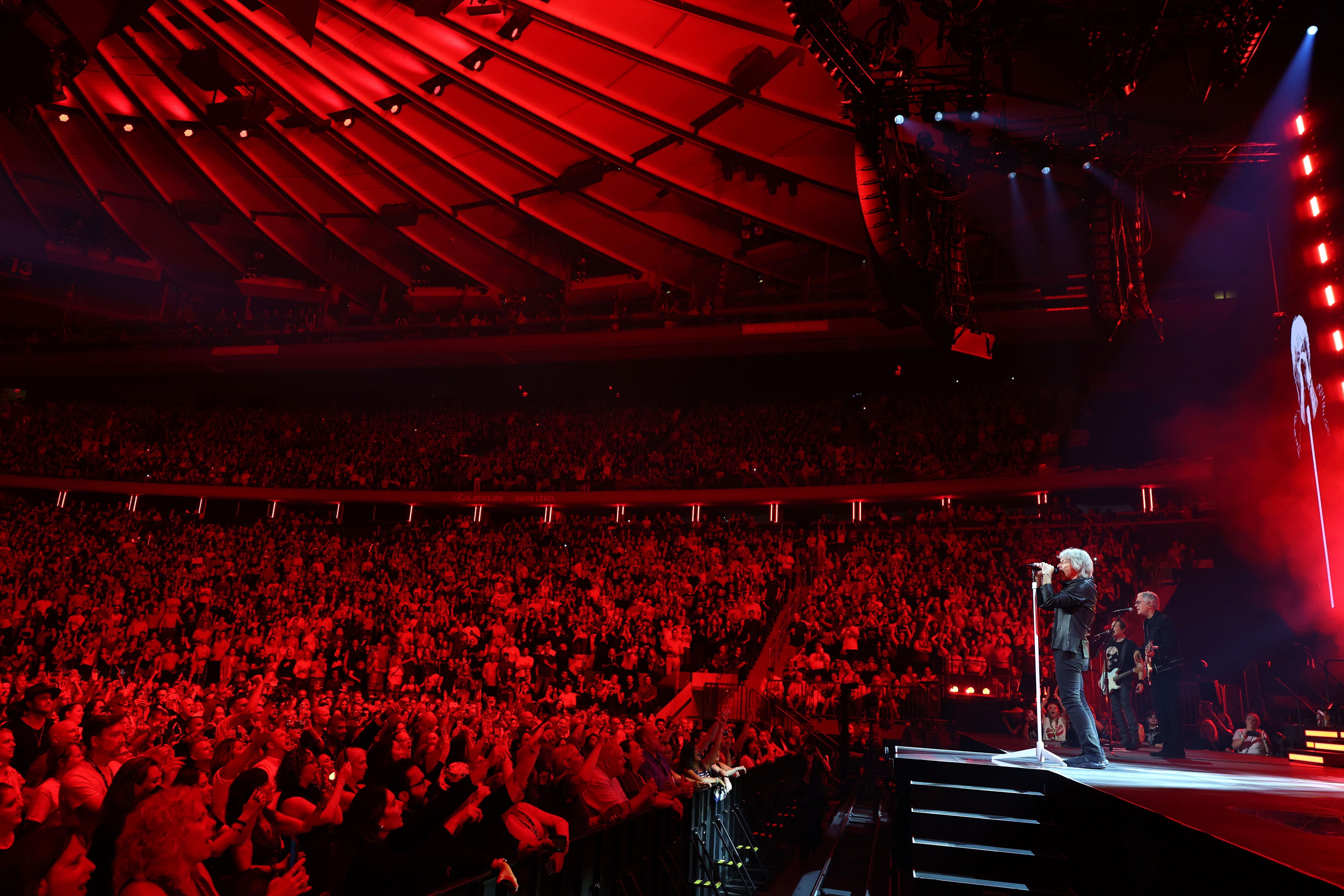 Konzert mit Band auf Bühne im roten Licht, große jubelnde Menschenmenge in einer Arena.
