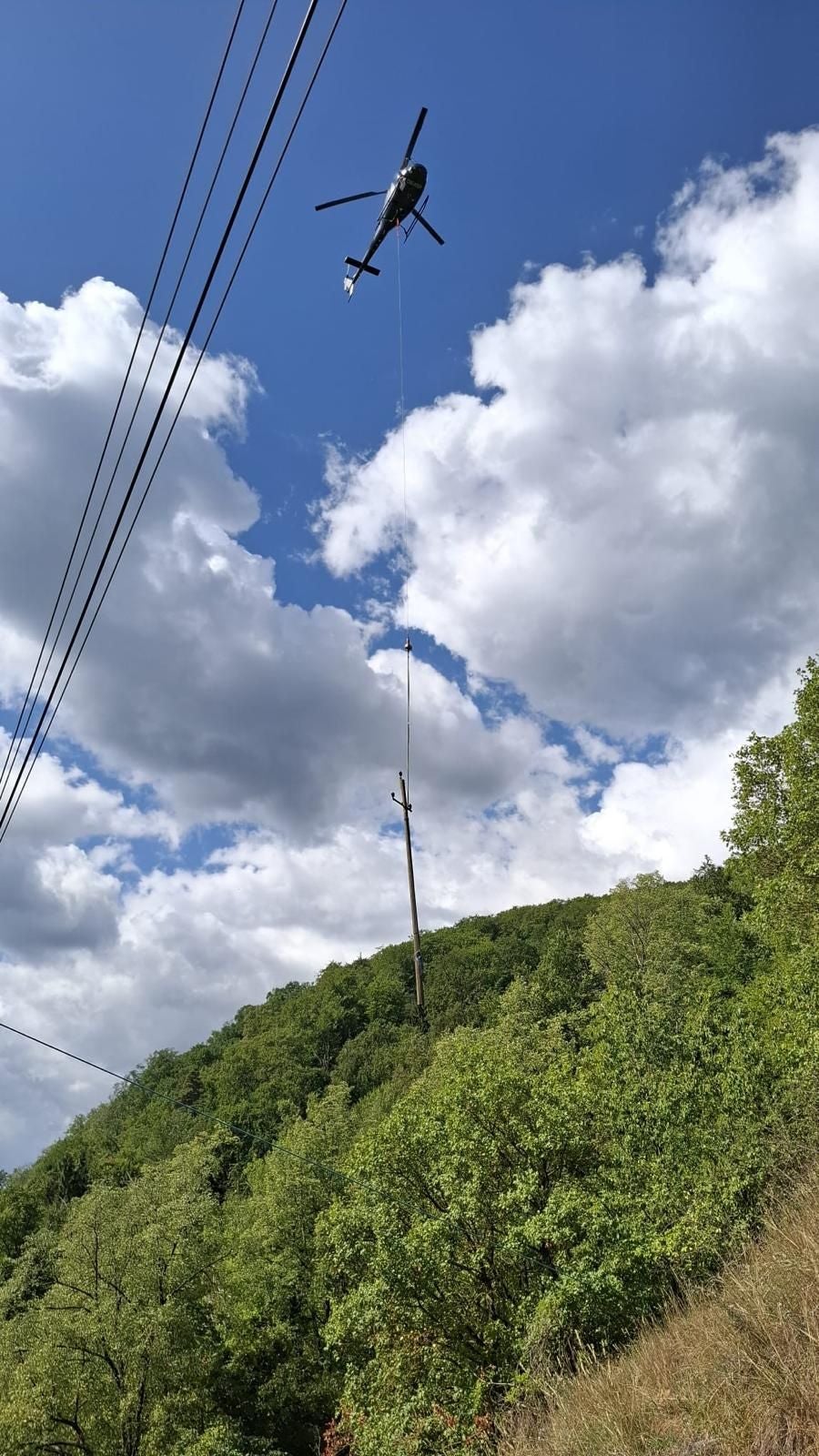 Ein Hubschrauber transportiert einen Strommast über einen bewaldeten Hügel bei blauem Himmel.