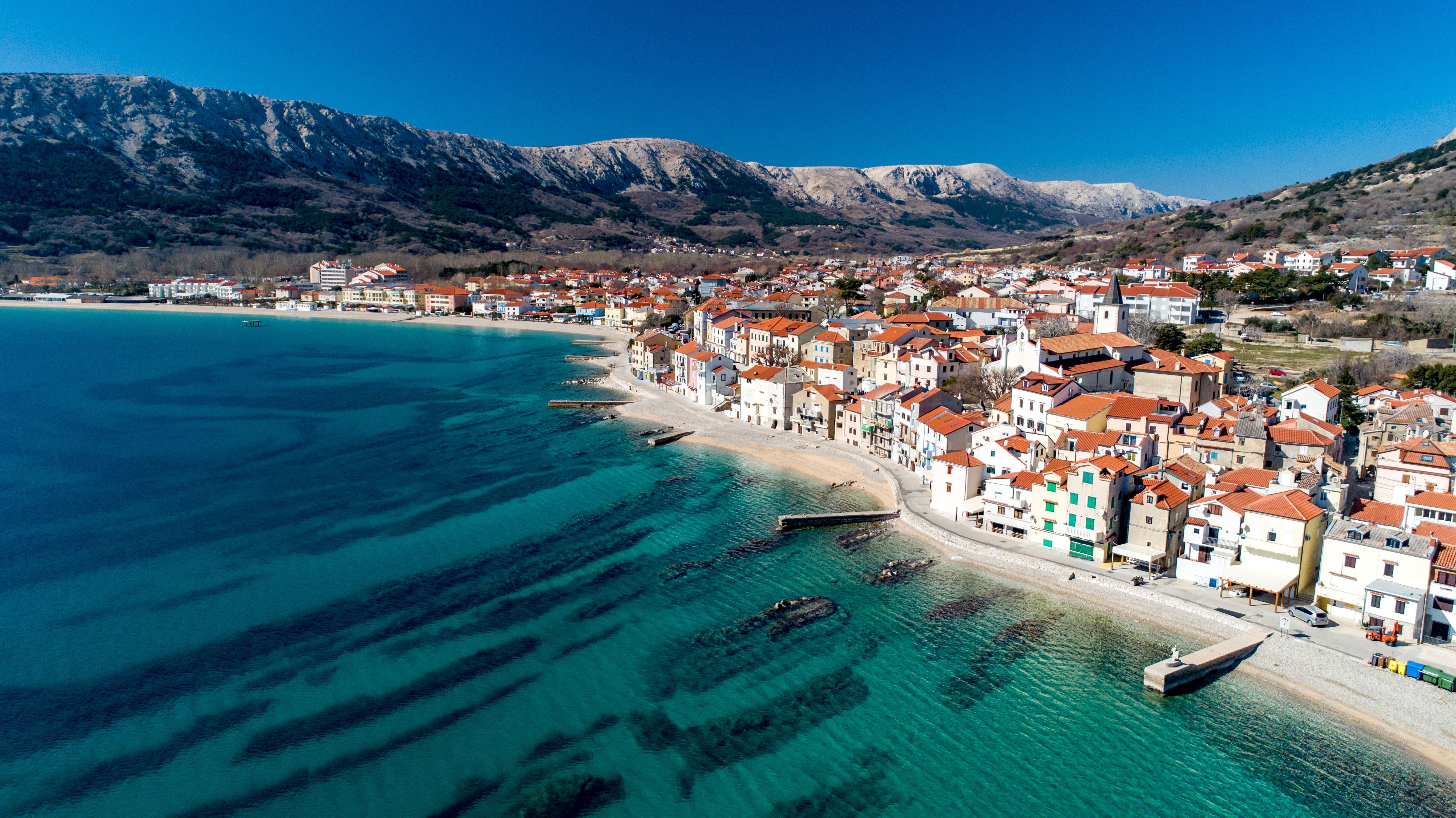 Luftaufnahme der Küstenstadt Baška auf der Insel Krk mit türkisblauem Meer und Bergen im Hintergrund.