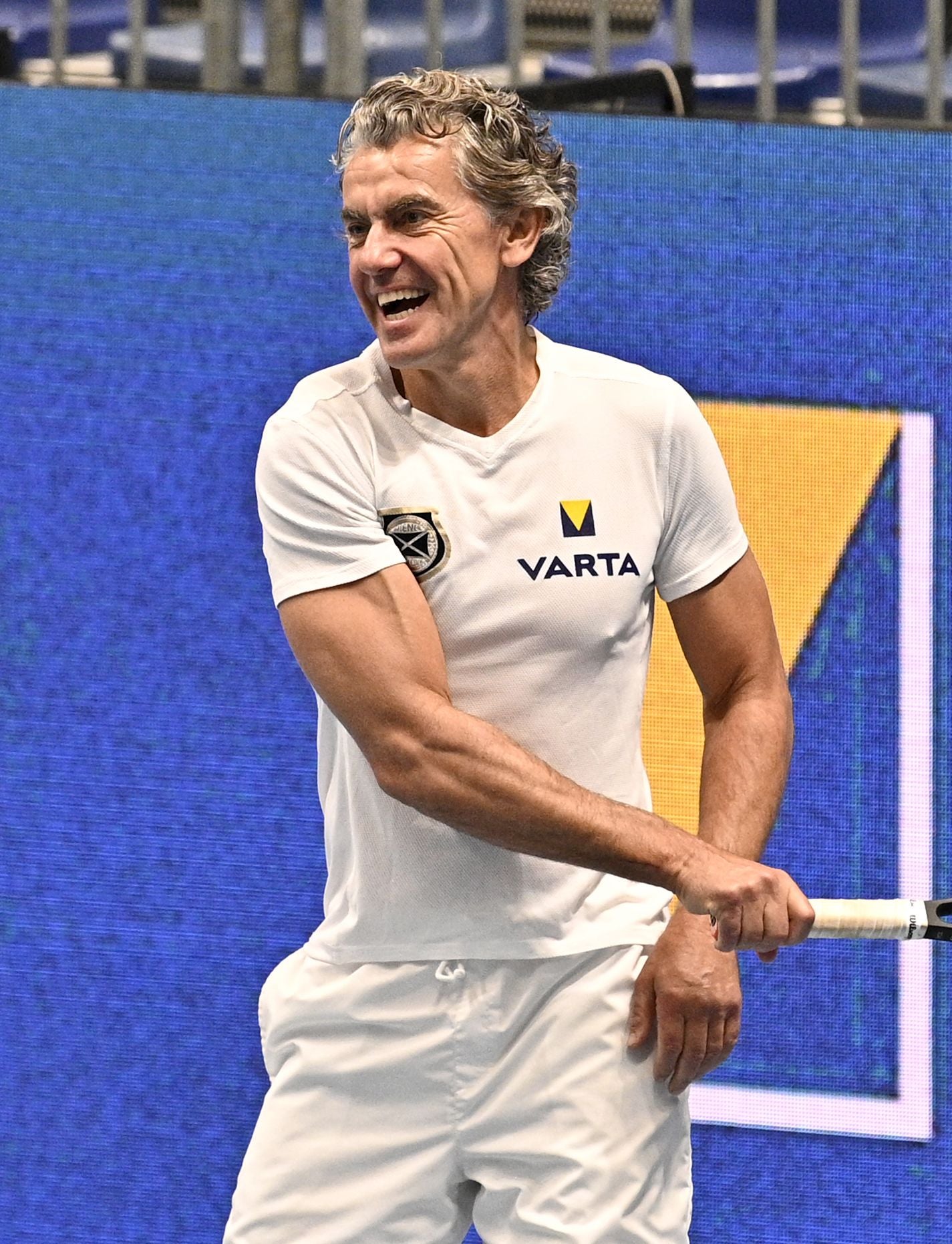 Ein Mann in weißem Sportoutfit hält einen Tennisschläger und lacht auf einem Tennisplatz.