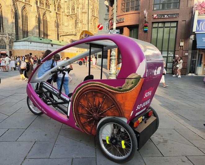Ein violettes Fahrradtaxi steht vor dem Stephansdom in Wien auf einem belebten Platz.