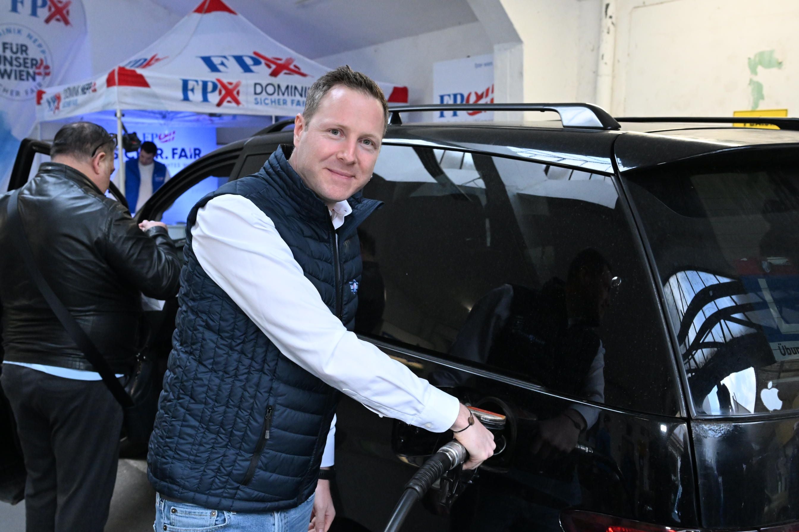 Ein Mann tankt ein Auto bei einer FPÖ-Spritaktion, im Hintergrund stehen weitere Personen.