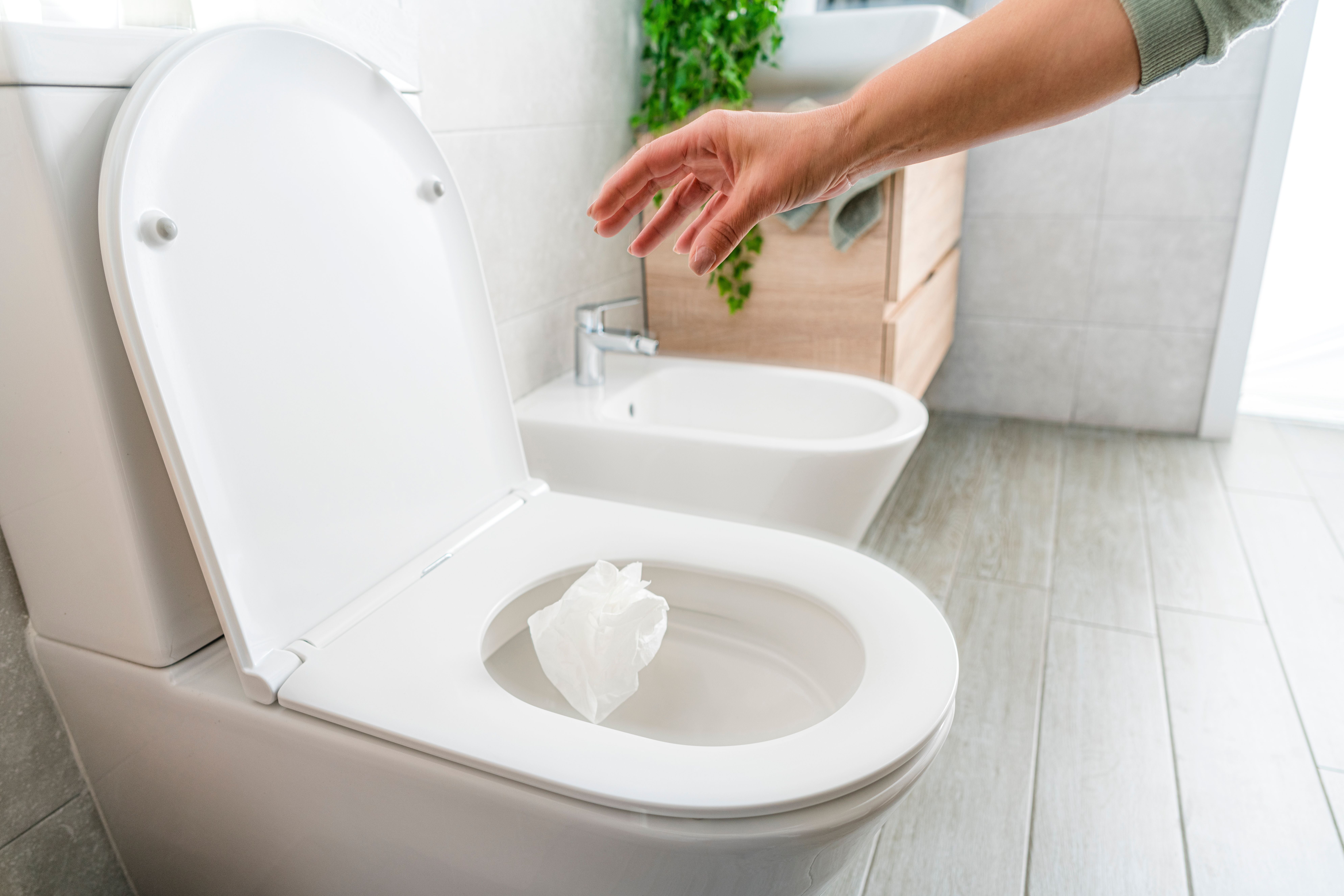 Hand wirft ein Stück Toilettenpapier in eine weiße Toilettenschüssel im Badezimmer.