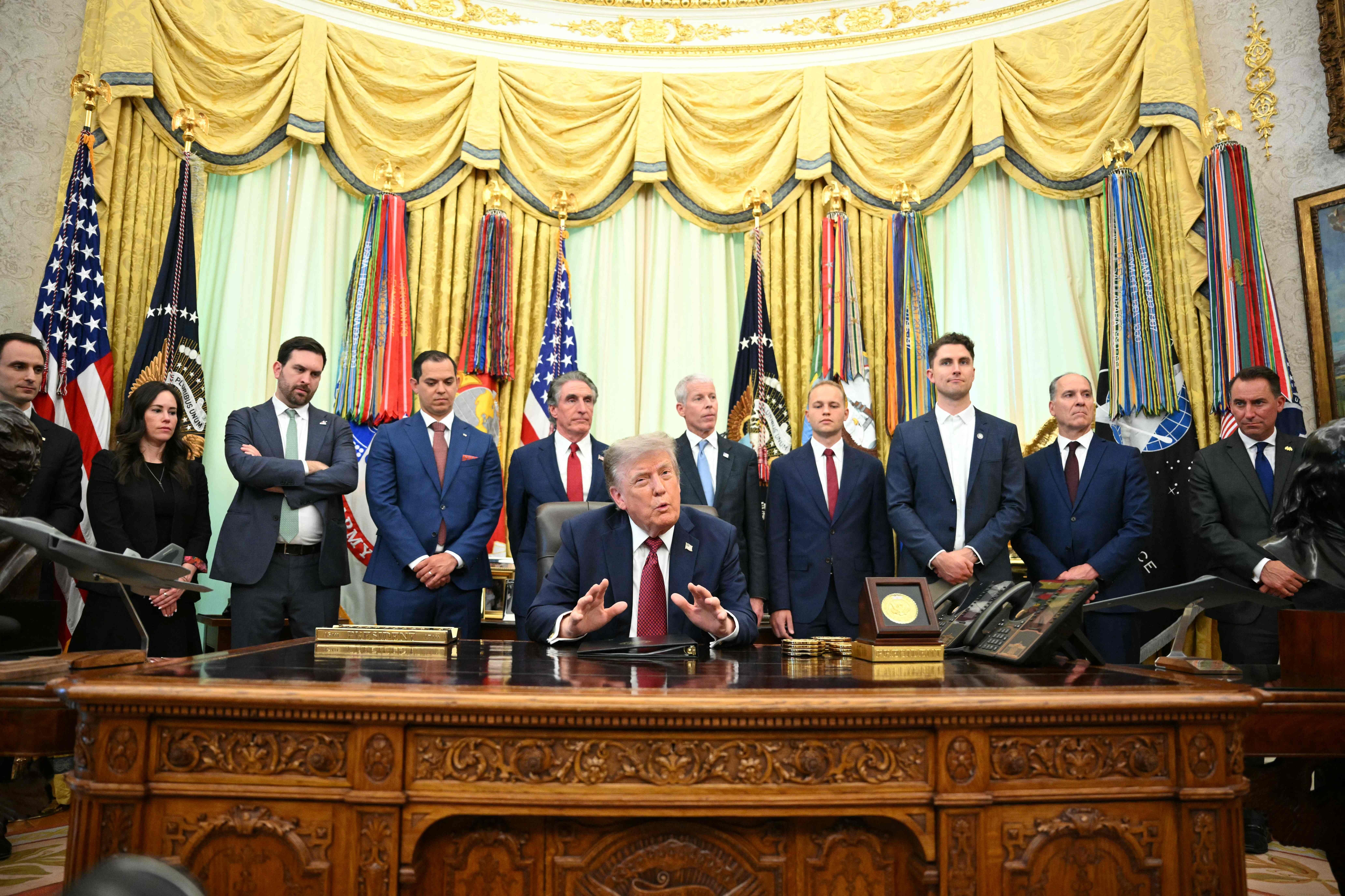 Donald Trump sitzt am Schreibtisch im Oval Office, umgeben von mehreren Menschen und US-Flaggen.