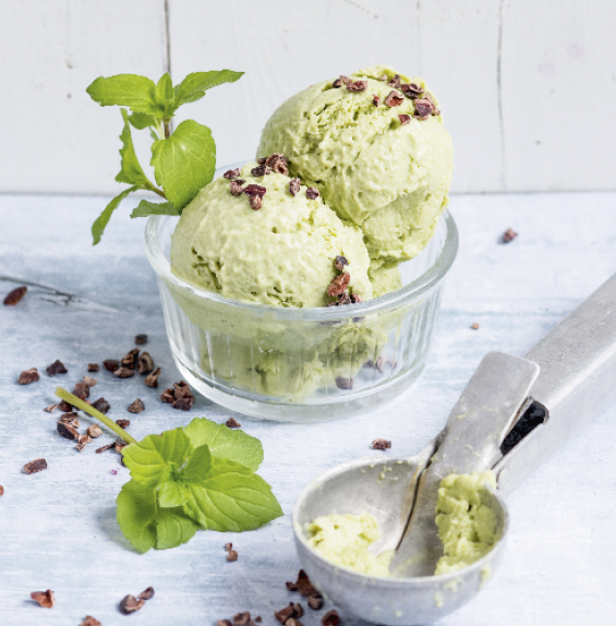 Zwei Kugeln grünes Eis mit Minze und Kakaostückchen in einem Glas, daneben ein Eislöffel.