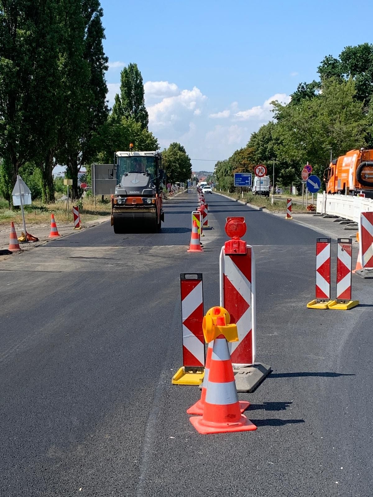 Straßenbaustelle mit Baufahrzeug, Verkehrshütchen und Absperrungen bei sonnigem Wetter.