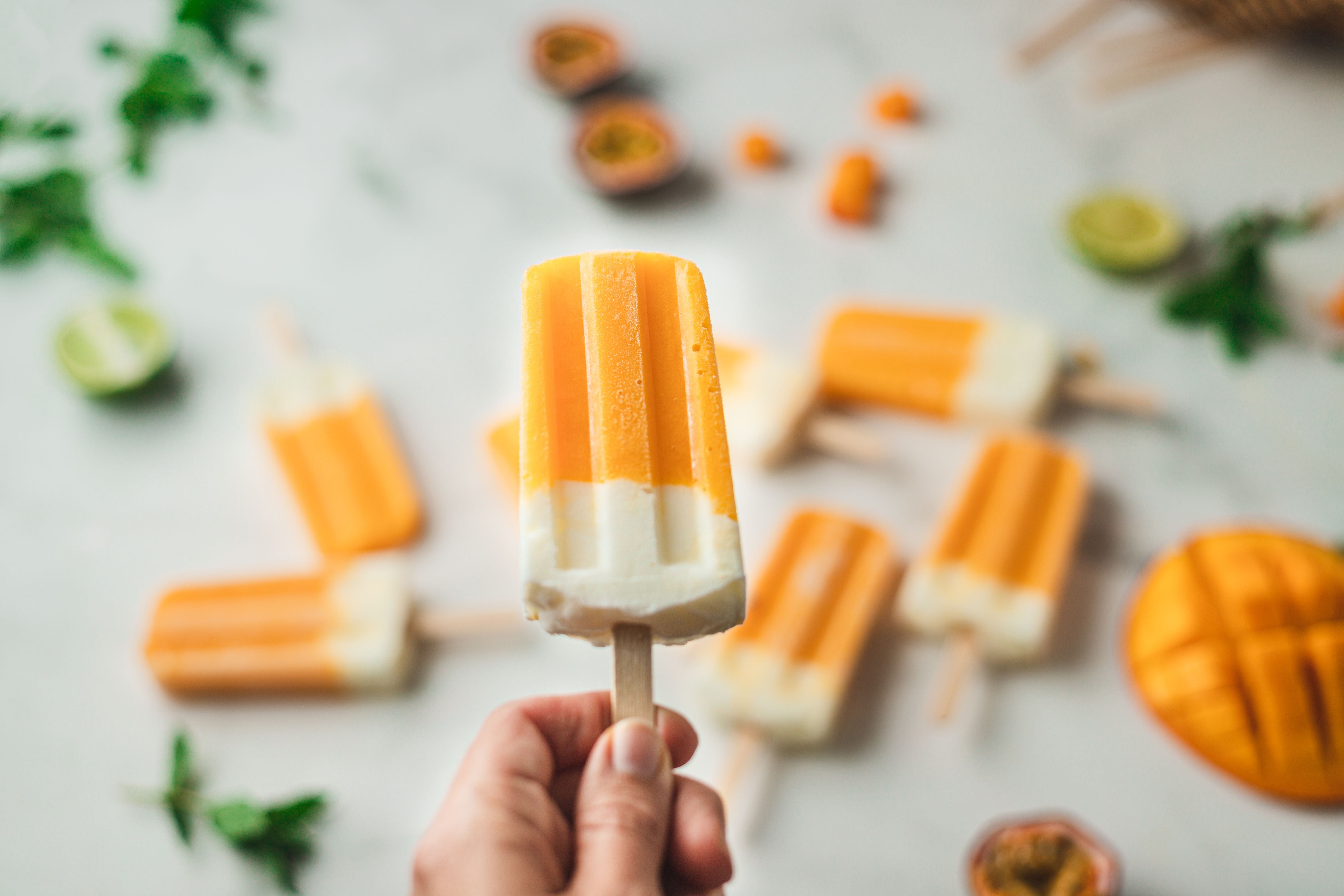 Hand hält ein Mango-Joghurt-Eis am Stiel, weitere Eis am Stiel und Zutaten im Hintergrund.