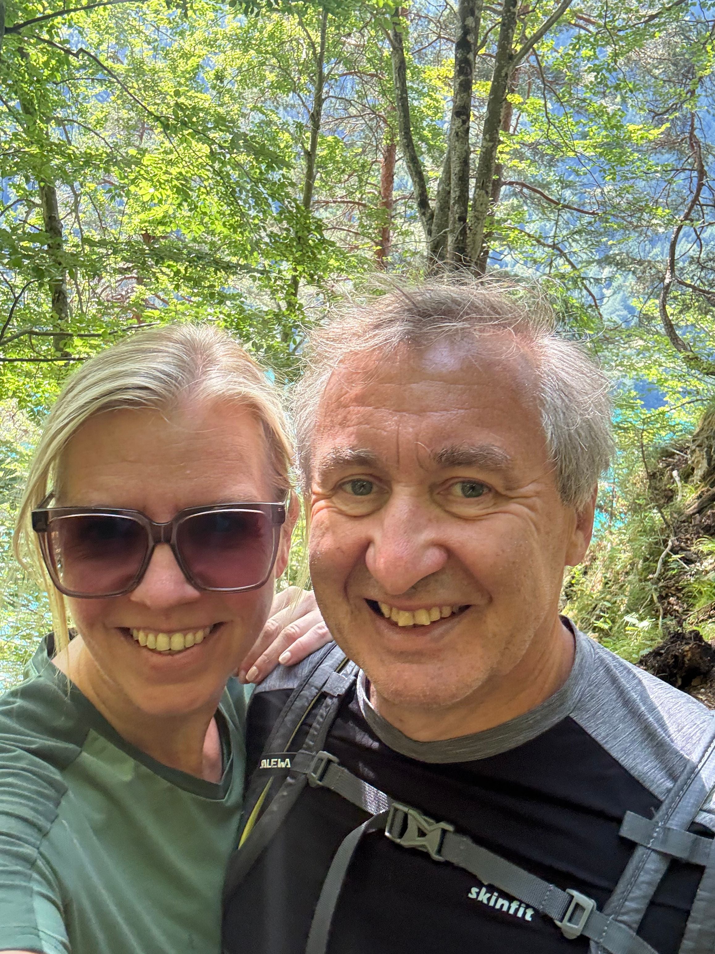 Zwei lachende Menschen machen ein Selfie beim Wandern im sonnigen Wald.