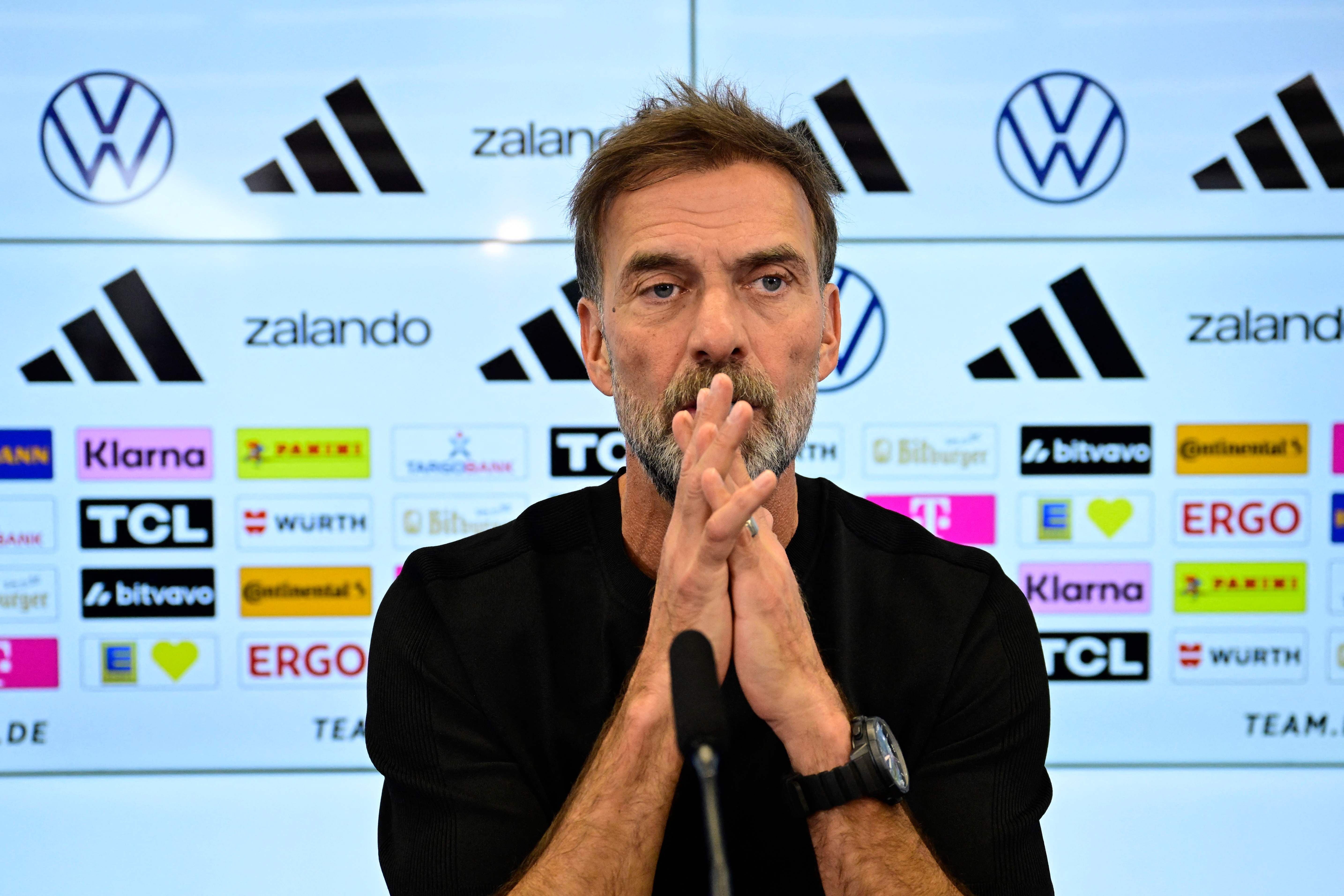Ein Mann mit Bart spricht nachdenklich bei einer Pressekonferenz vor Sponsoren-Logos.