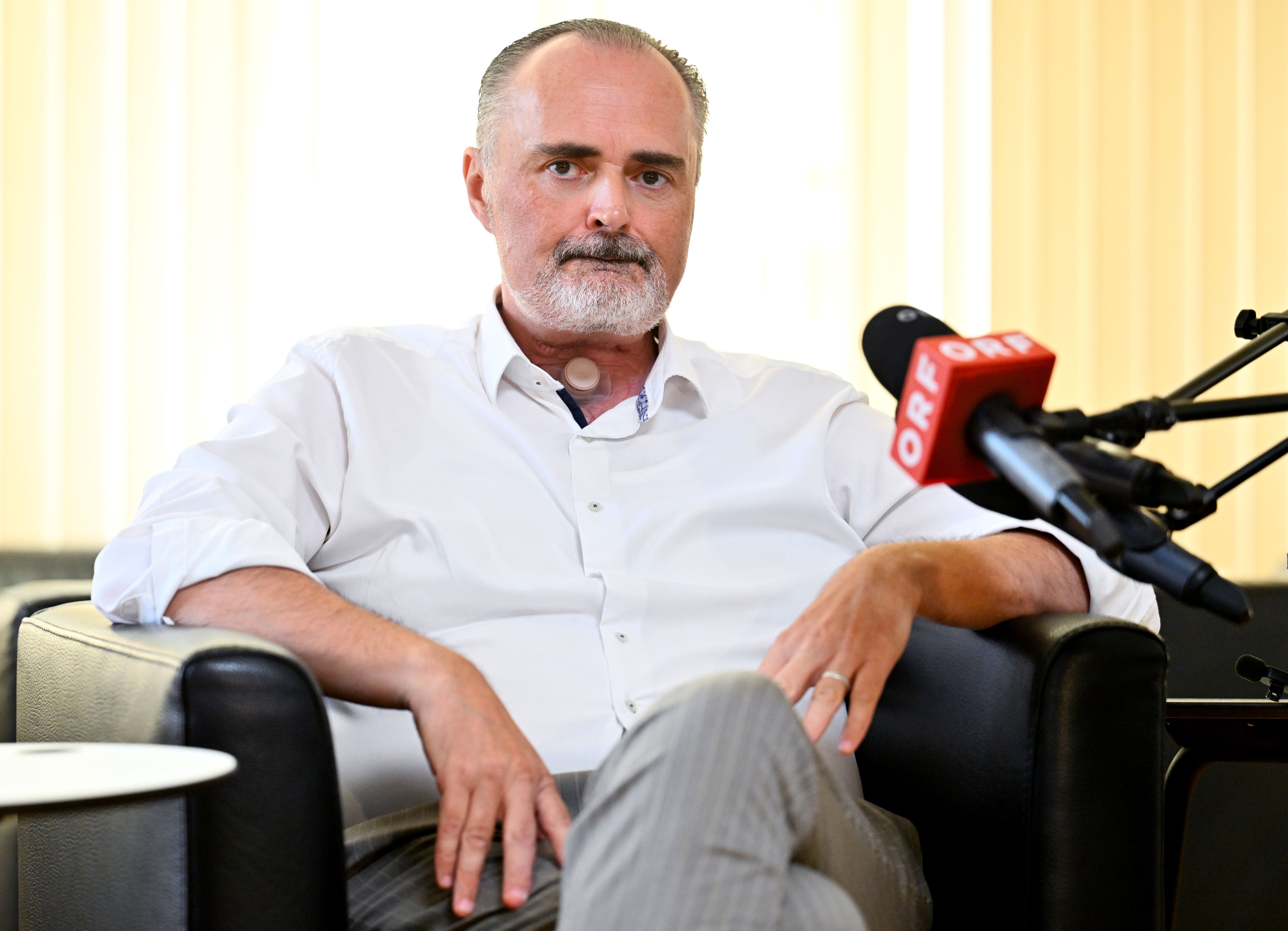 Mann im weißen Hemd sitzt bei einer Pressekonferenz, Mikrofone vor ihm, ernster Ausdruck.