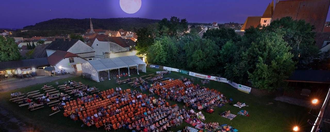 Menschen sitzen abends auf Stühlen und Decken bei einem Open-Air-Kino vor einer Stadt-Kulisse.