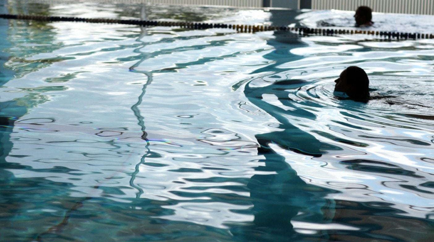 Zwei Personen schwimmen in einem Hallenbad mit ruhiger Wasseroberfläche.