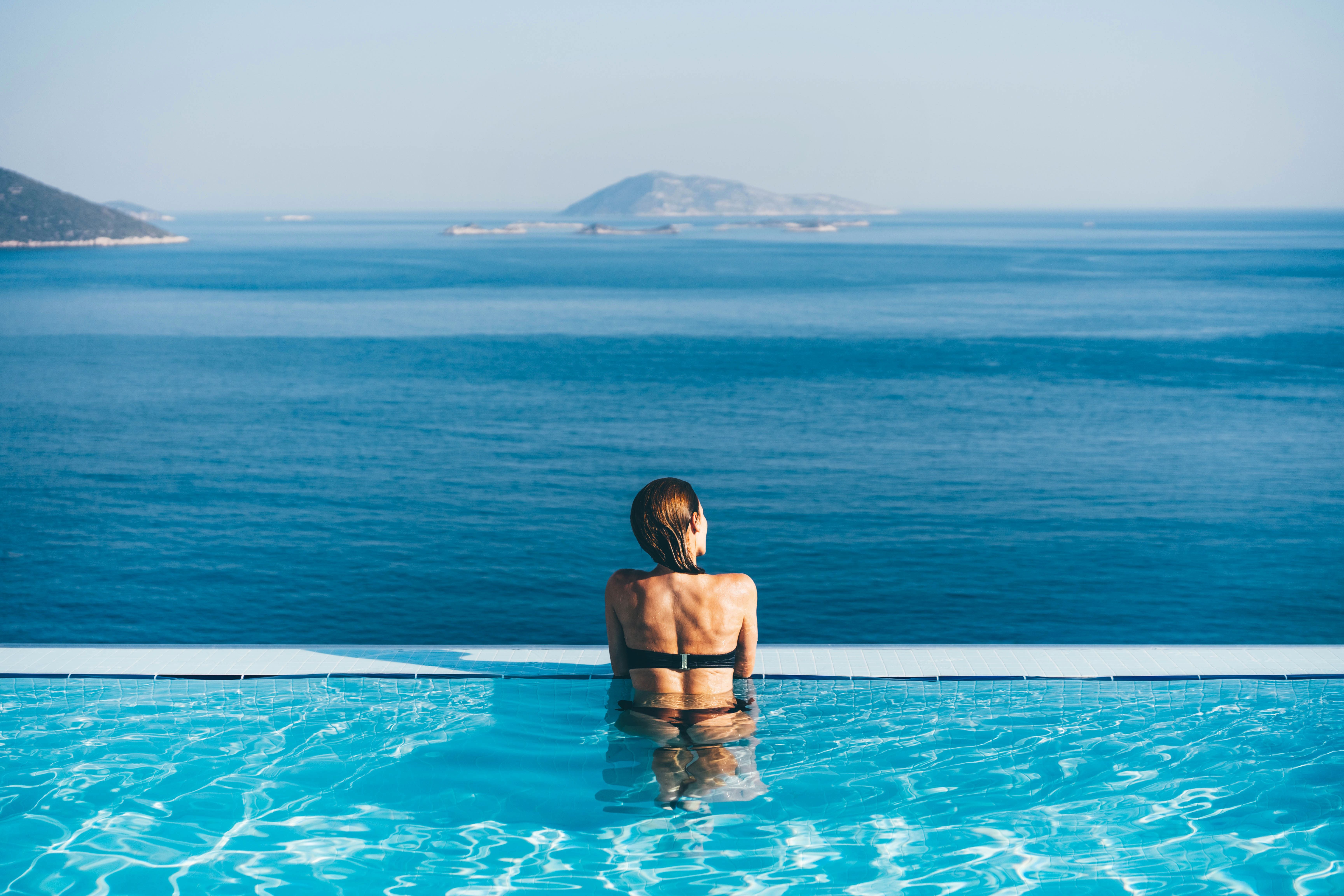 Frau sitzt im Infinity-Pool und blickt auf das weite blaue Meer mit einer Insel im Hintergrund.