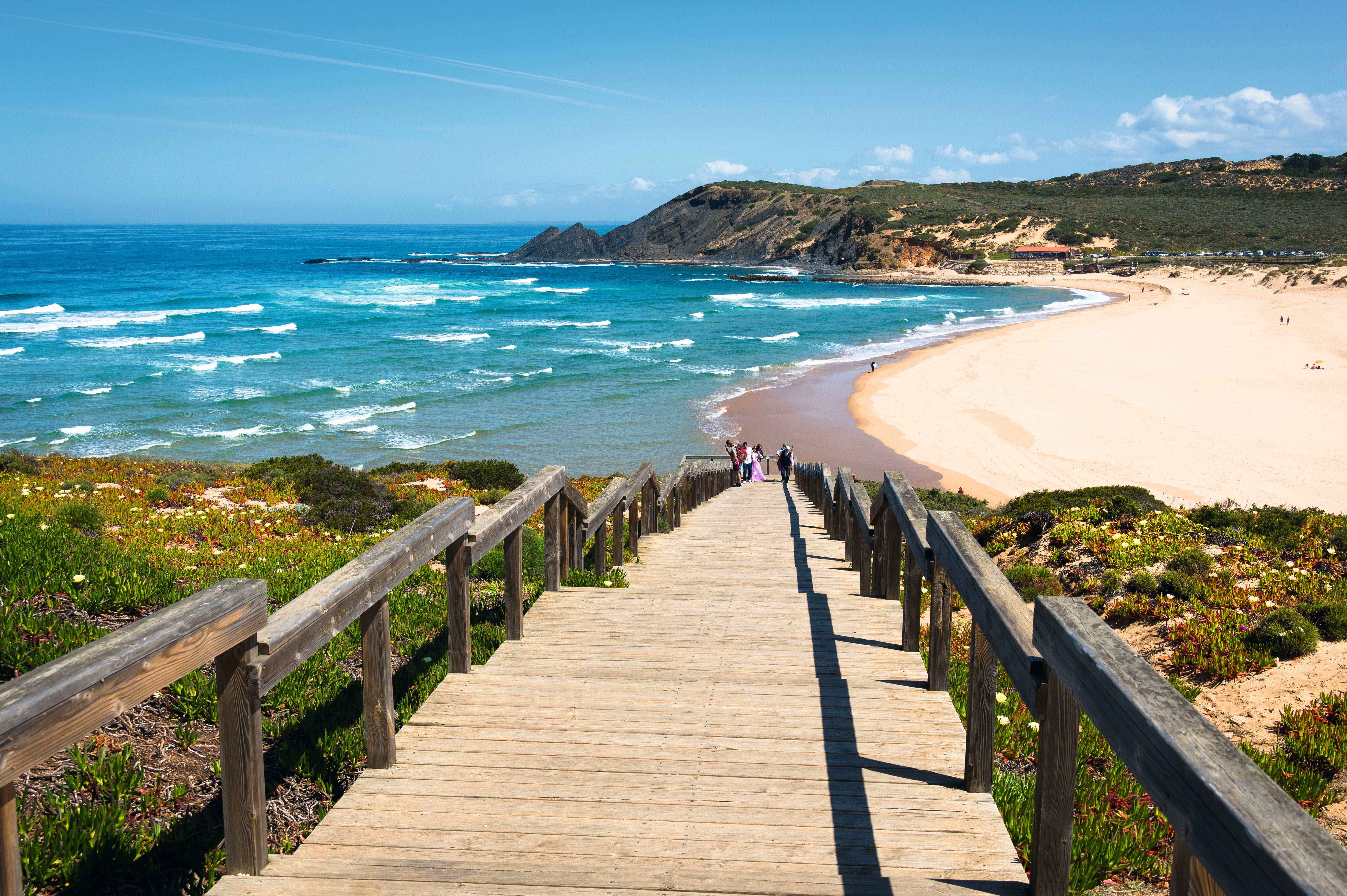 Holztreppe führt zu einem Sandstrand mit türkisfarbenem Meer und grünen Klippen im Hintergrund.