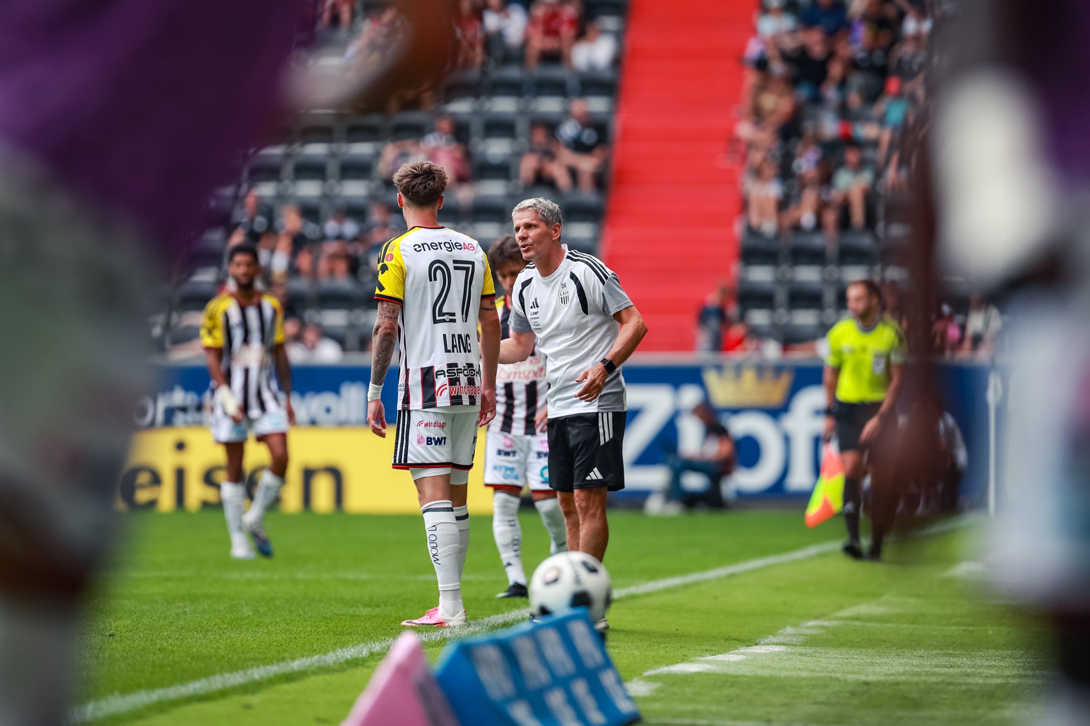 Ein Fußballtrainer im grauen Trainingsshirt spricht an der Seitenlinie auf einen Spieler im schwarz-weiß gestreiften Trikot mit der Rückennummer 27 und dem Namen Lang ein.