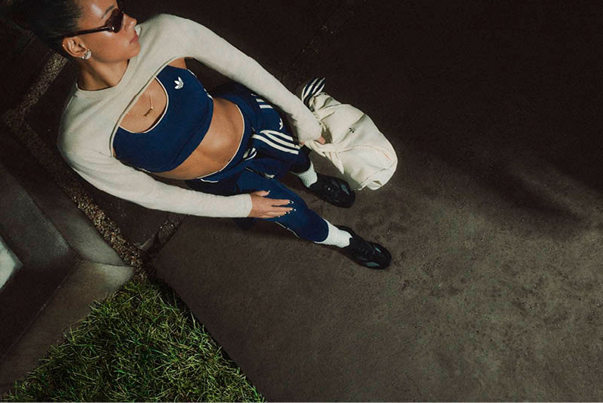 Frau in adidas Sportkleidung und Sonnenbrille hält eine helle Tasche auf einem Gehweg.