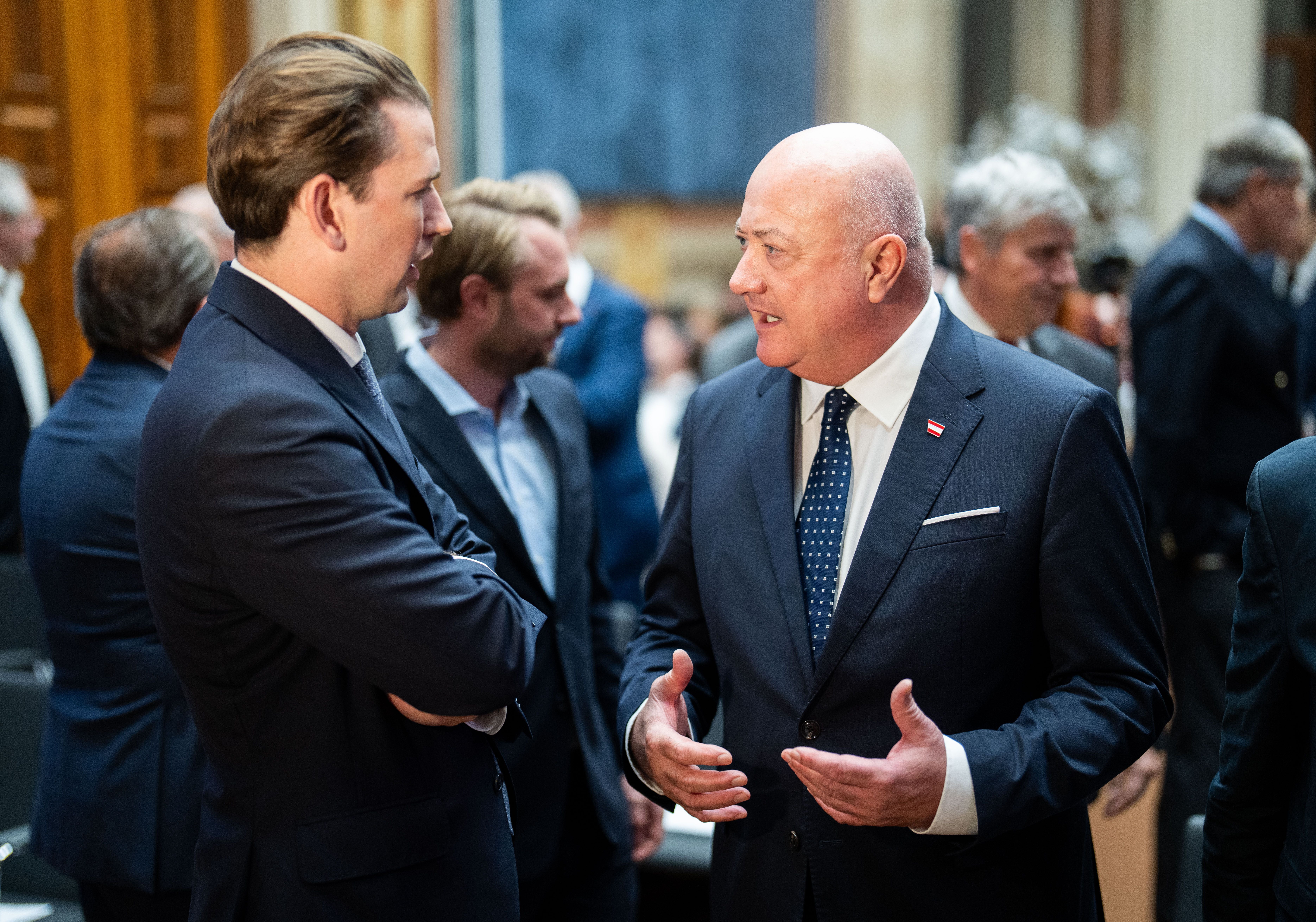 Zwei Männer im Anzug unterhalten sich bei einer Veranstaltung in einem festlichen Saal.