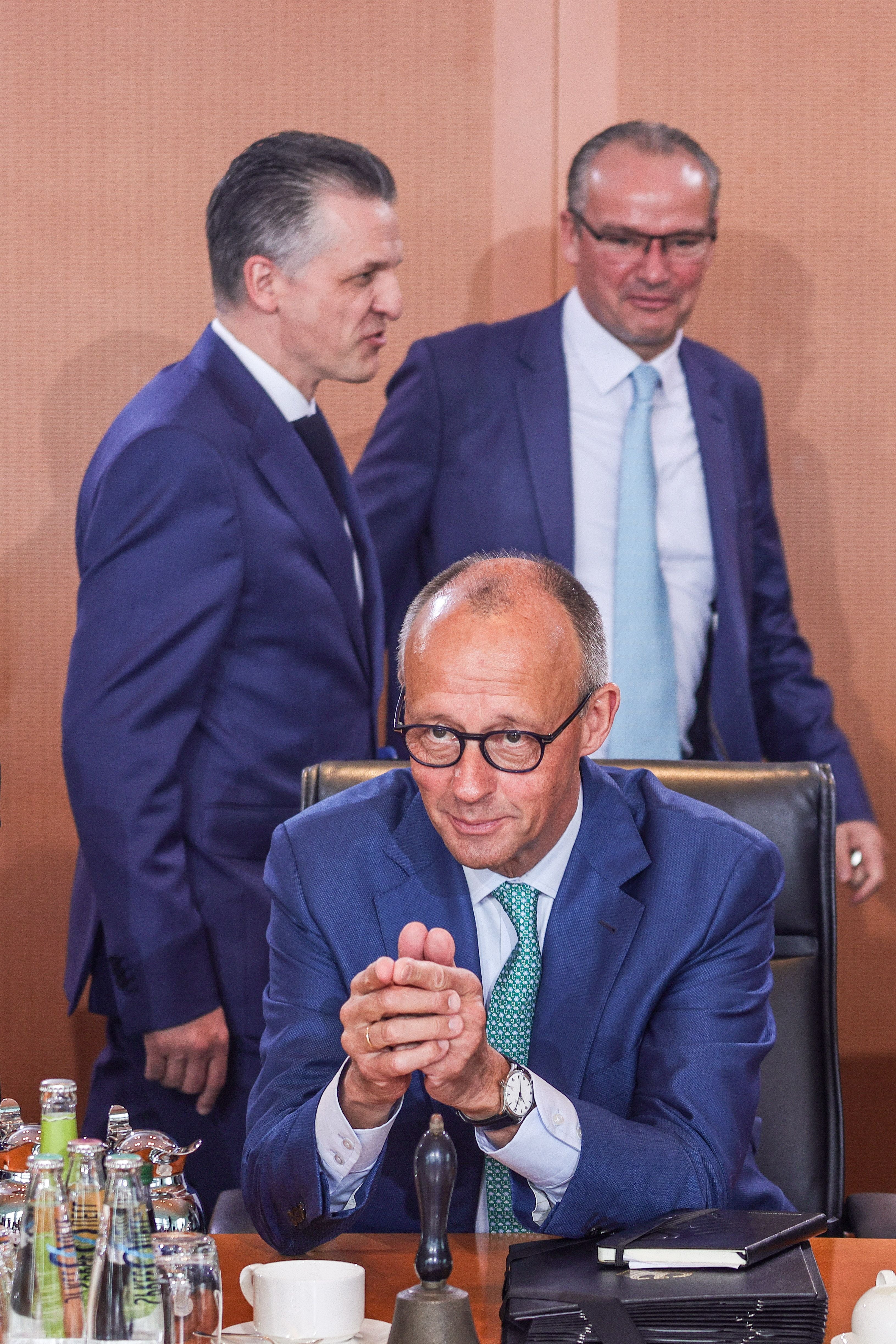 Drei Männer im Anzug bei einer Kabinettssitzung, einer sitzt, zwei stehen im Hintergrund.