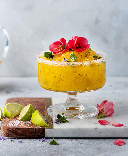 Gelbes Eis in einem Glas, garniert mit rosa Blüten und Minze, daneben Limettenstücke.