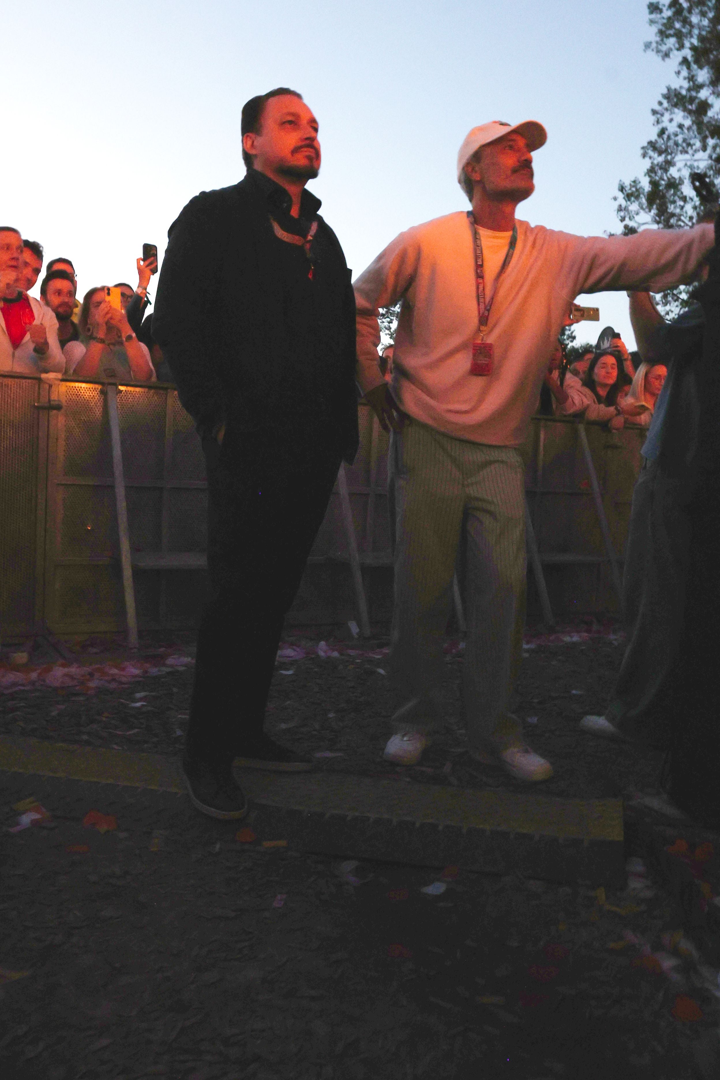 Zwei Männer stehen vor einer Festivalbühne, Menschenmenge im Hintergrund bei Abenddämmerung.