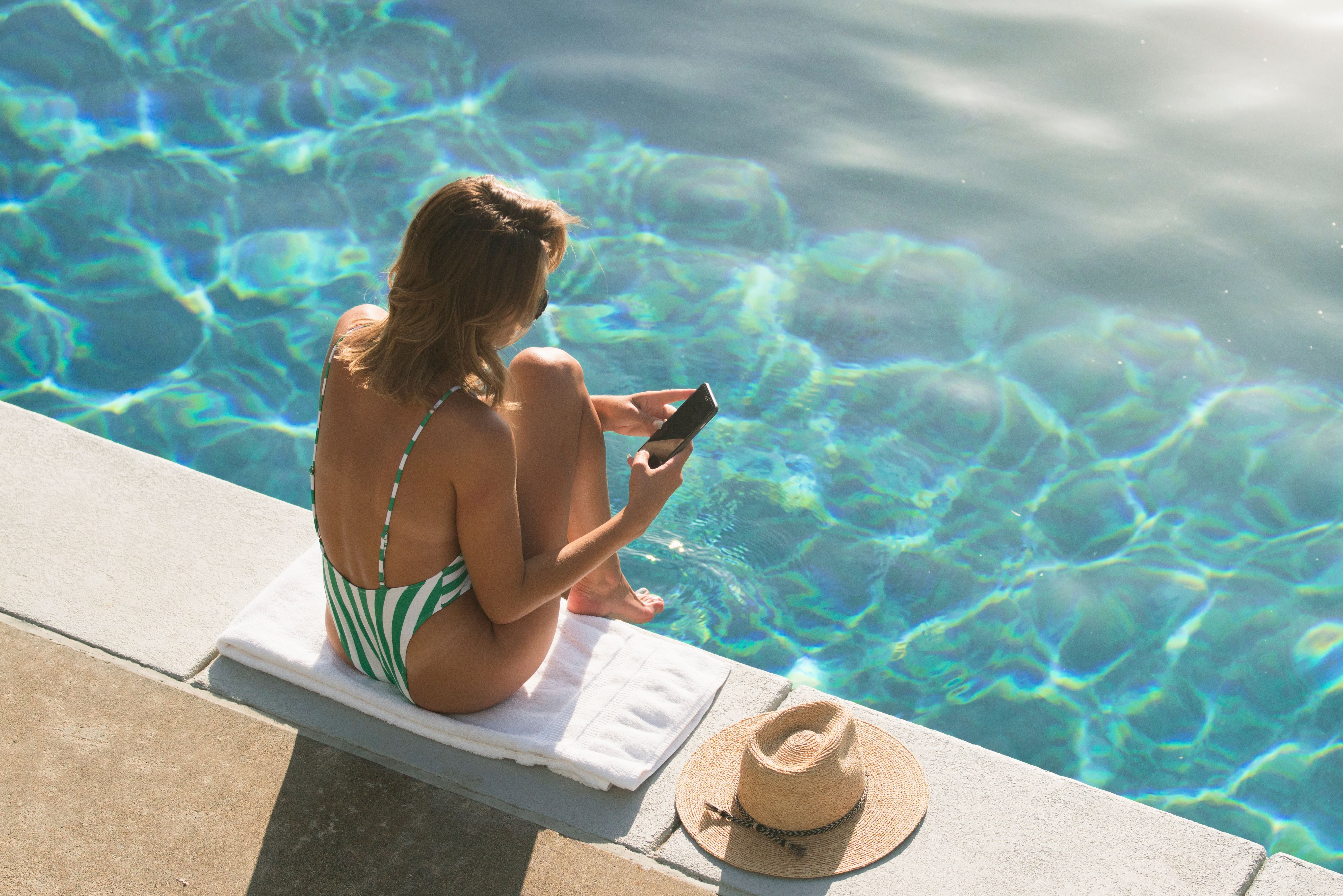 Frau im Badeanzug sitzt am Pool, schaut auf ihr Handy, ein Strohhut liegt neben ihr.