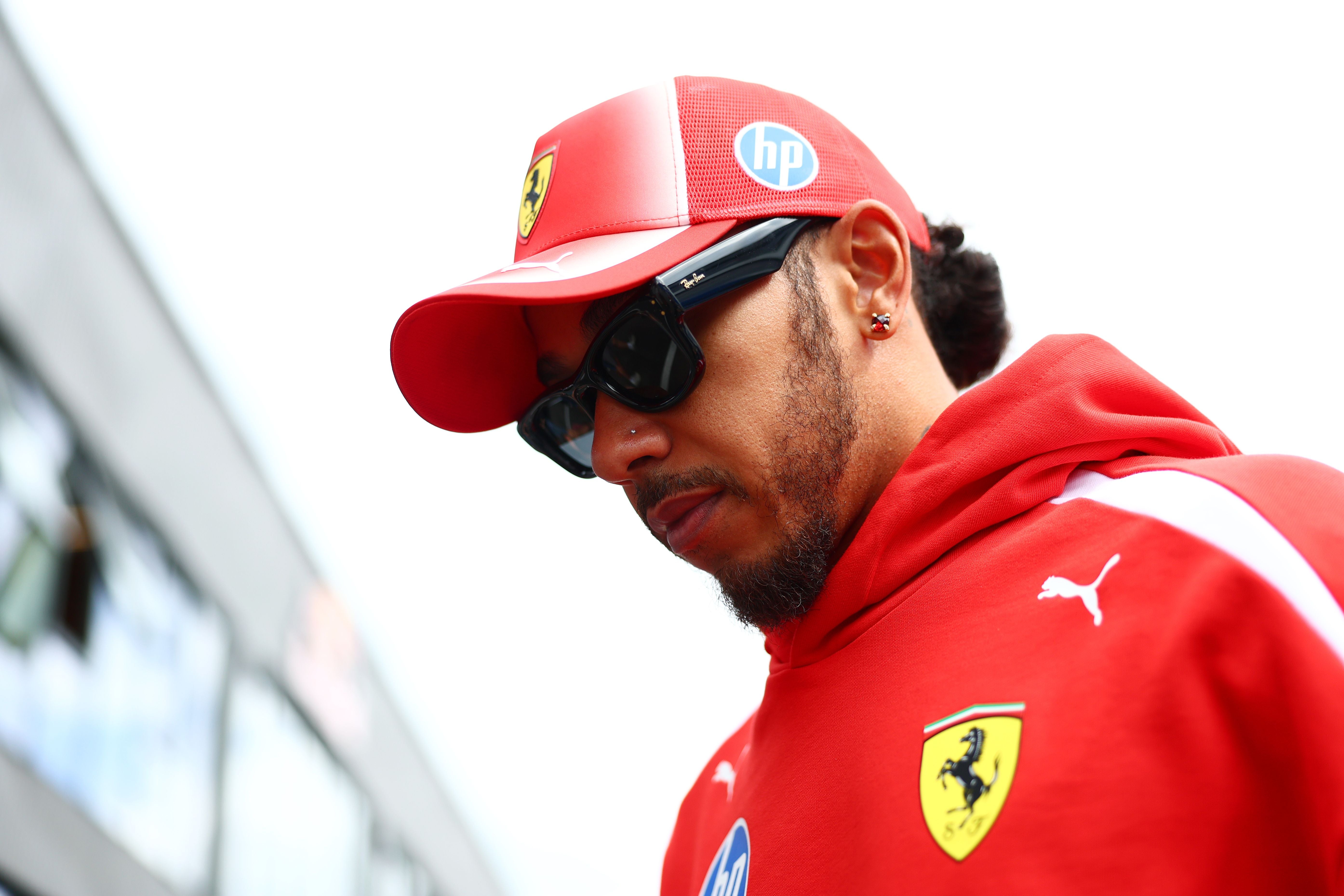 Lewis Hamilton im roten Ferrari-Teamoutfit mit roter Kappe und Sonnenbrille blickt im Freien gesenkt nach unten.