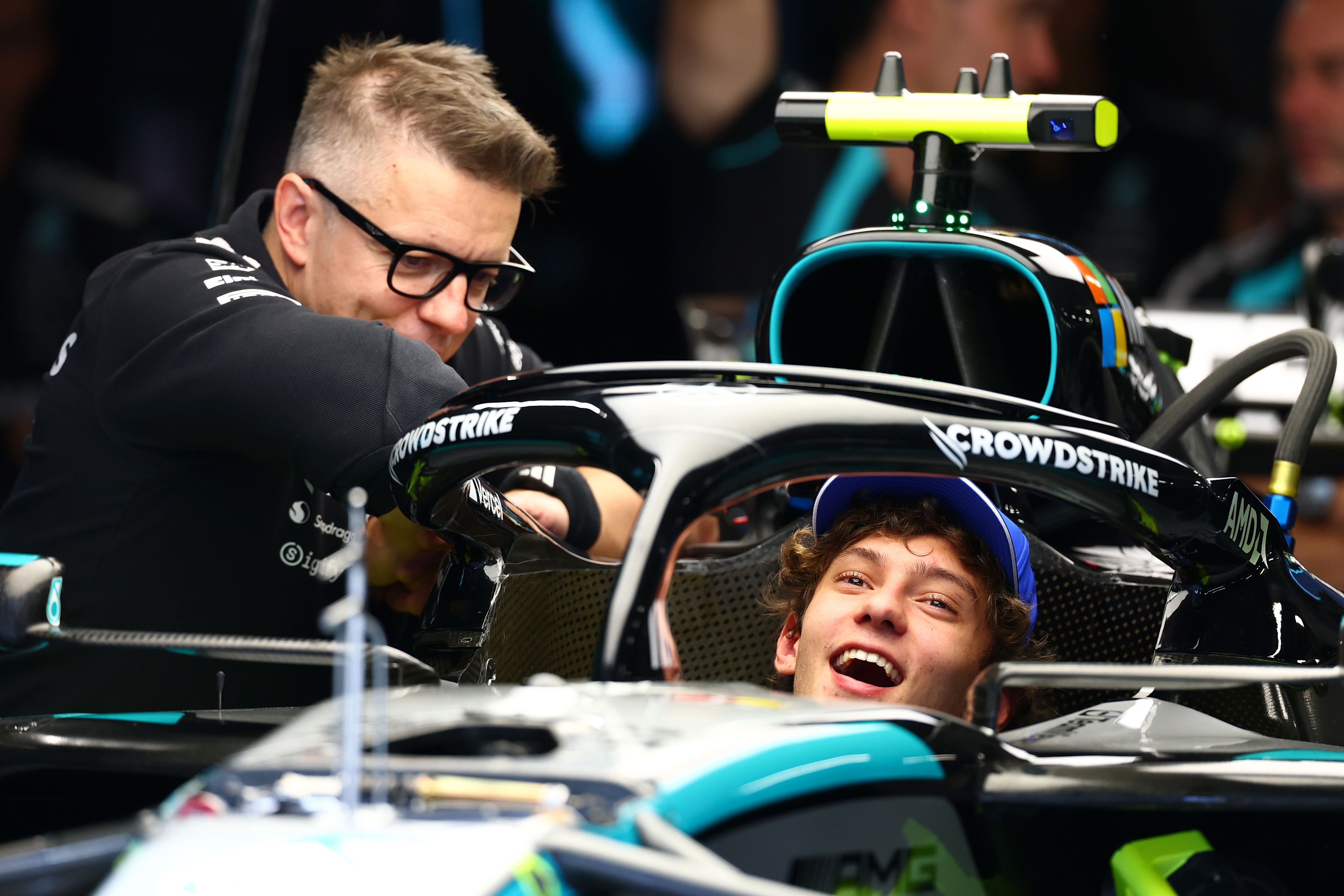 Kimi Antonelli sitzt lächelnd im Cockpit eines Mercedes-Formel-1-Wagens, während ein Teammitglied mit Brille von oben auf ihn herab blickt.