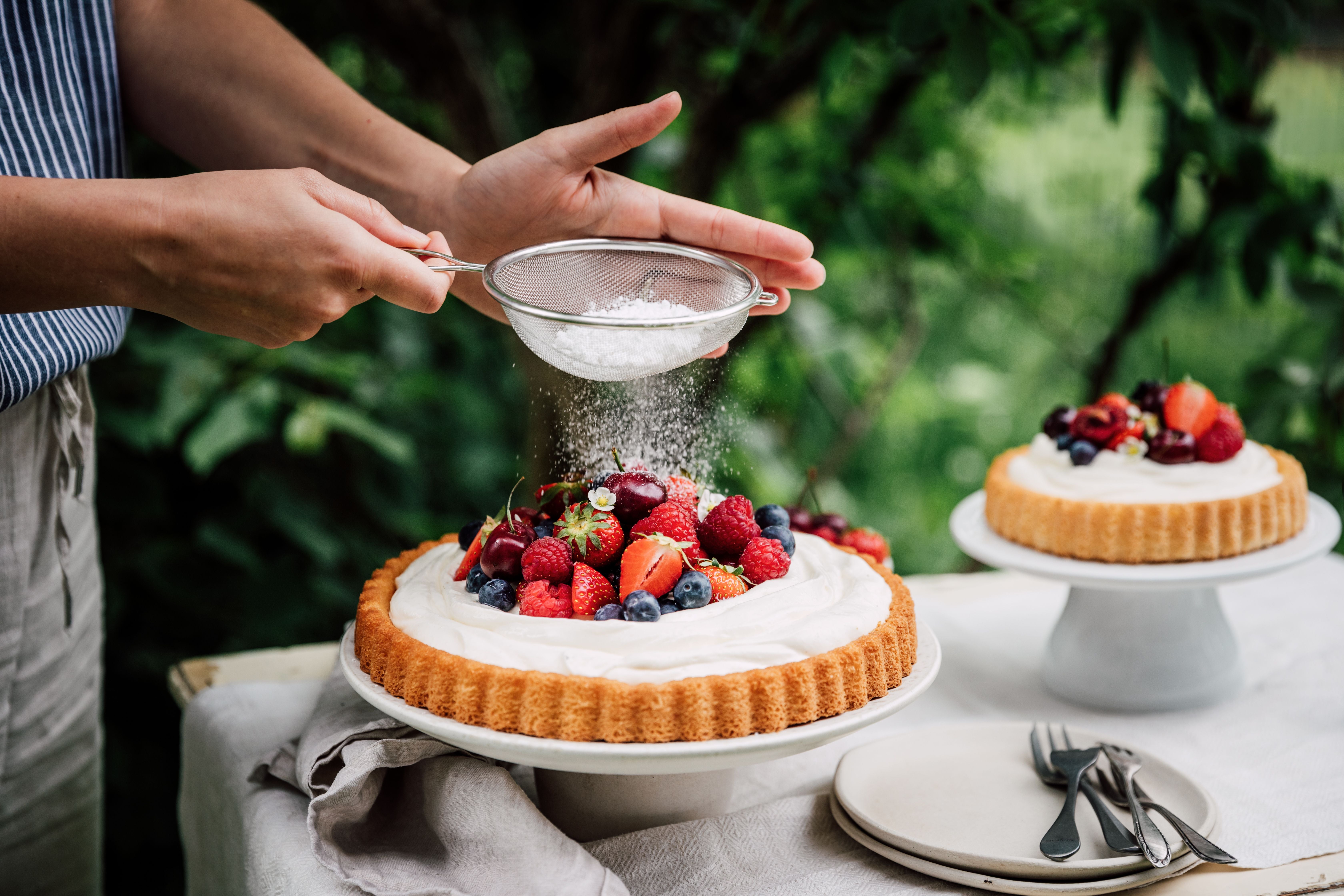 Hände bestäuben eine Beeren-Torte mit Puderzucker im Garten auf einem gedeckten Tisch.
