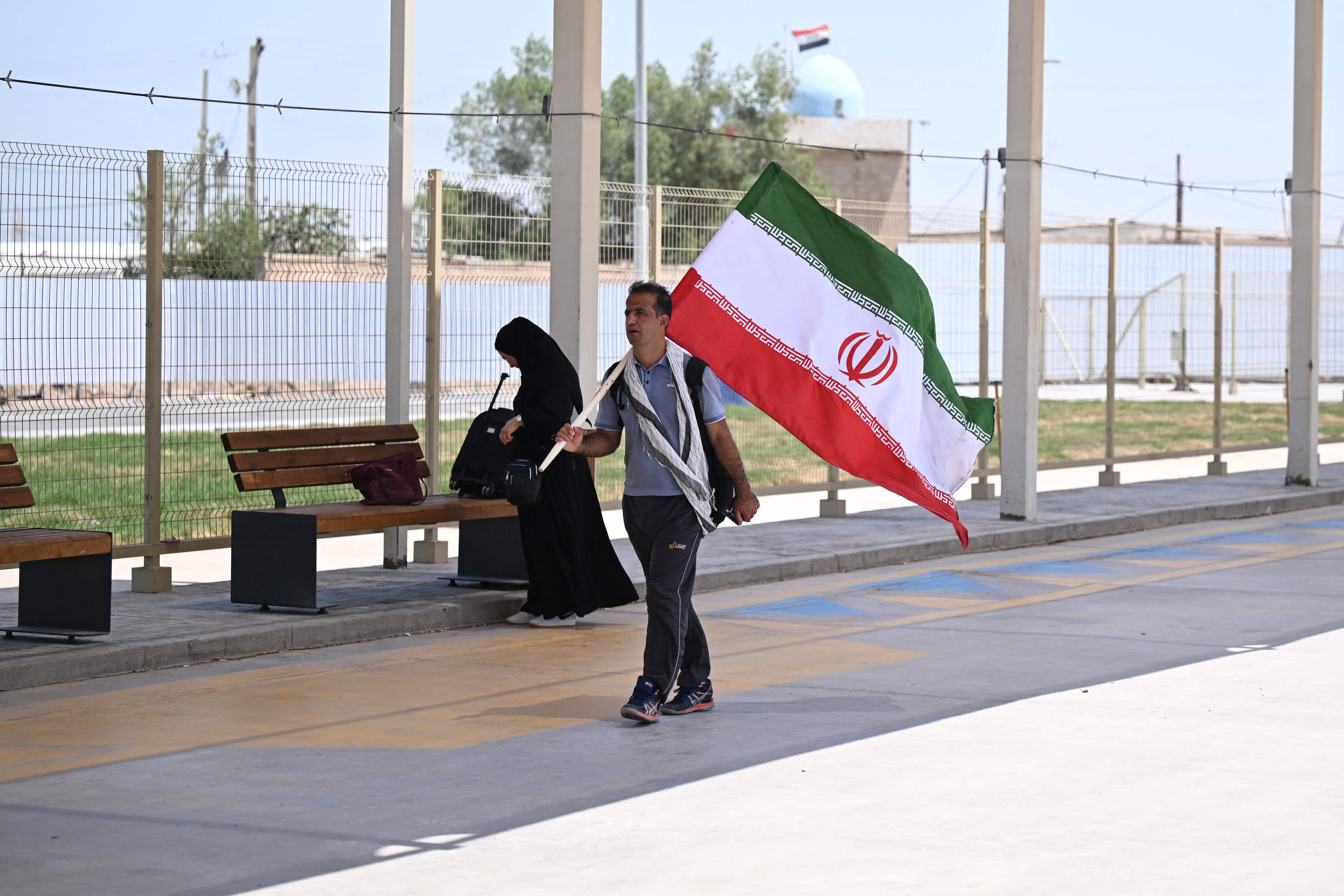 Ein Mann trägt die iranische Flagge an einem Grenzübergang; im Hintergrund sitzt eine Frau.