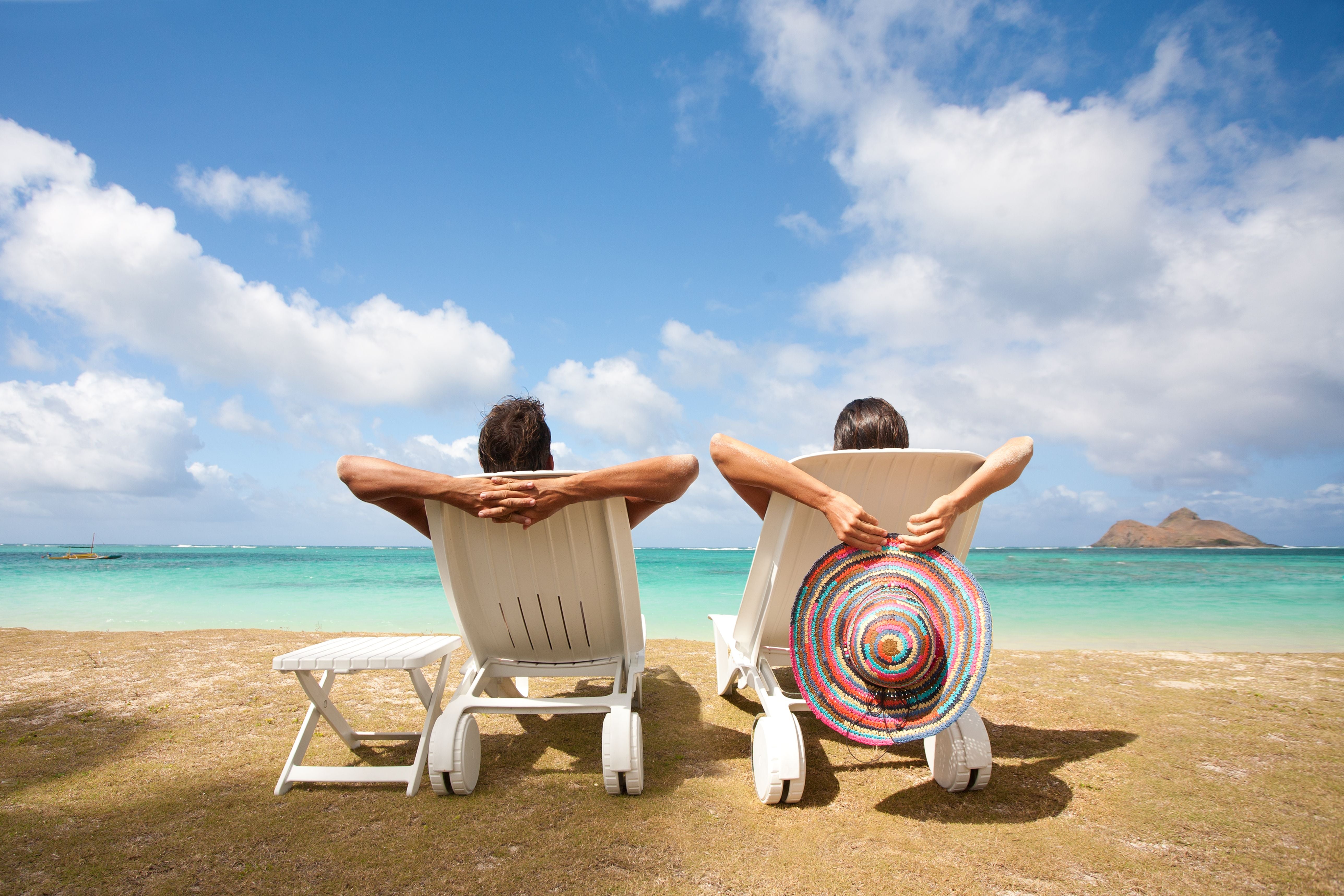 Pärchen entspannt in Liegestühlen am Sandstrand mit türkisfarbenem Meer und blauem Himmel.