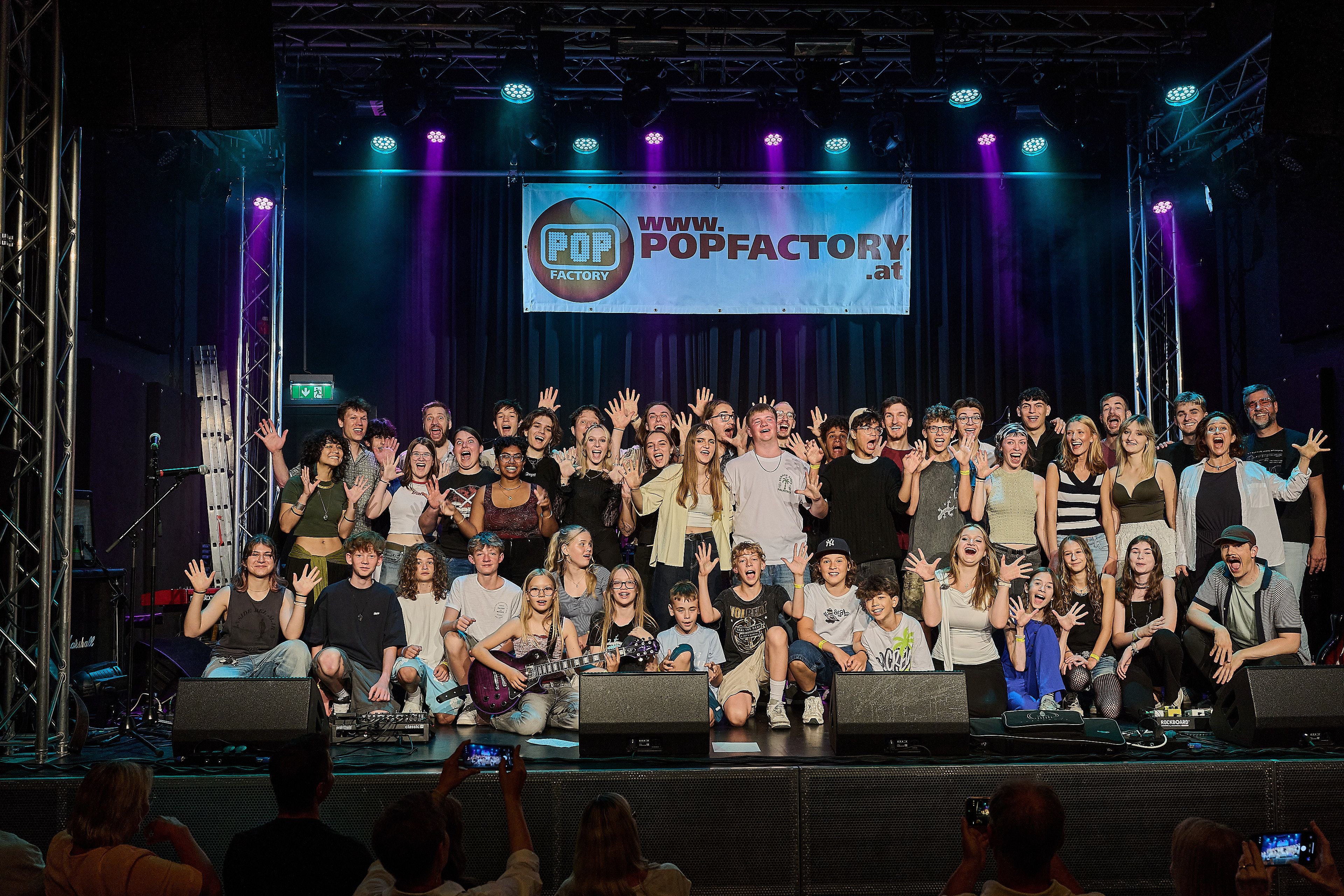 Eine große Gruppe junger Menschen posiert lachend auf einer Bühnen mit „Popfactory“-Banner.