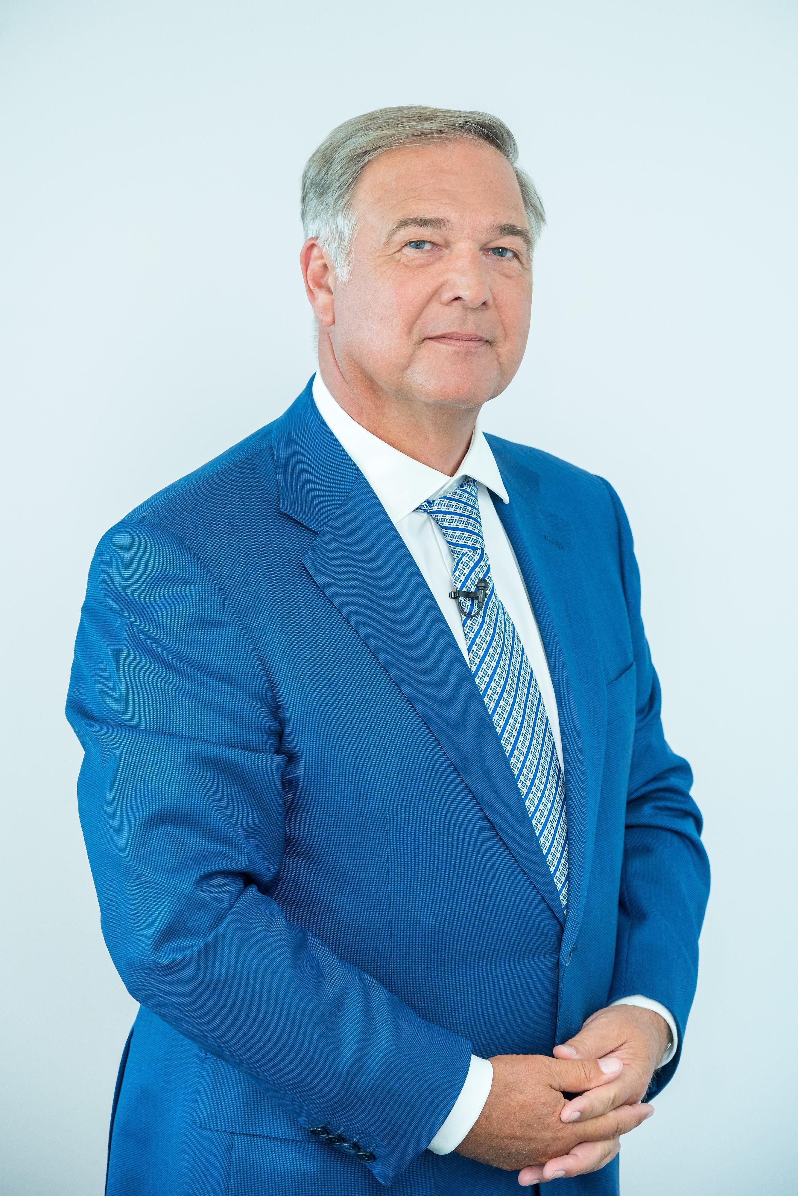 Ein Mann im blauen Anzug mit Krawatte steht vor hellem Hintergrund und blickt in die Kamera.
