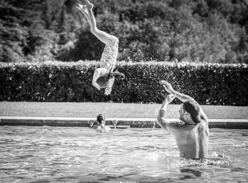 Ein Mann wirft ein Kind beim Spielen im Schwimmbad in die Luft, beide haben Spaß.