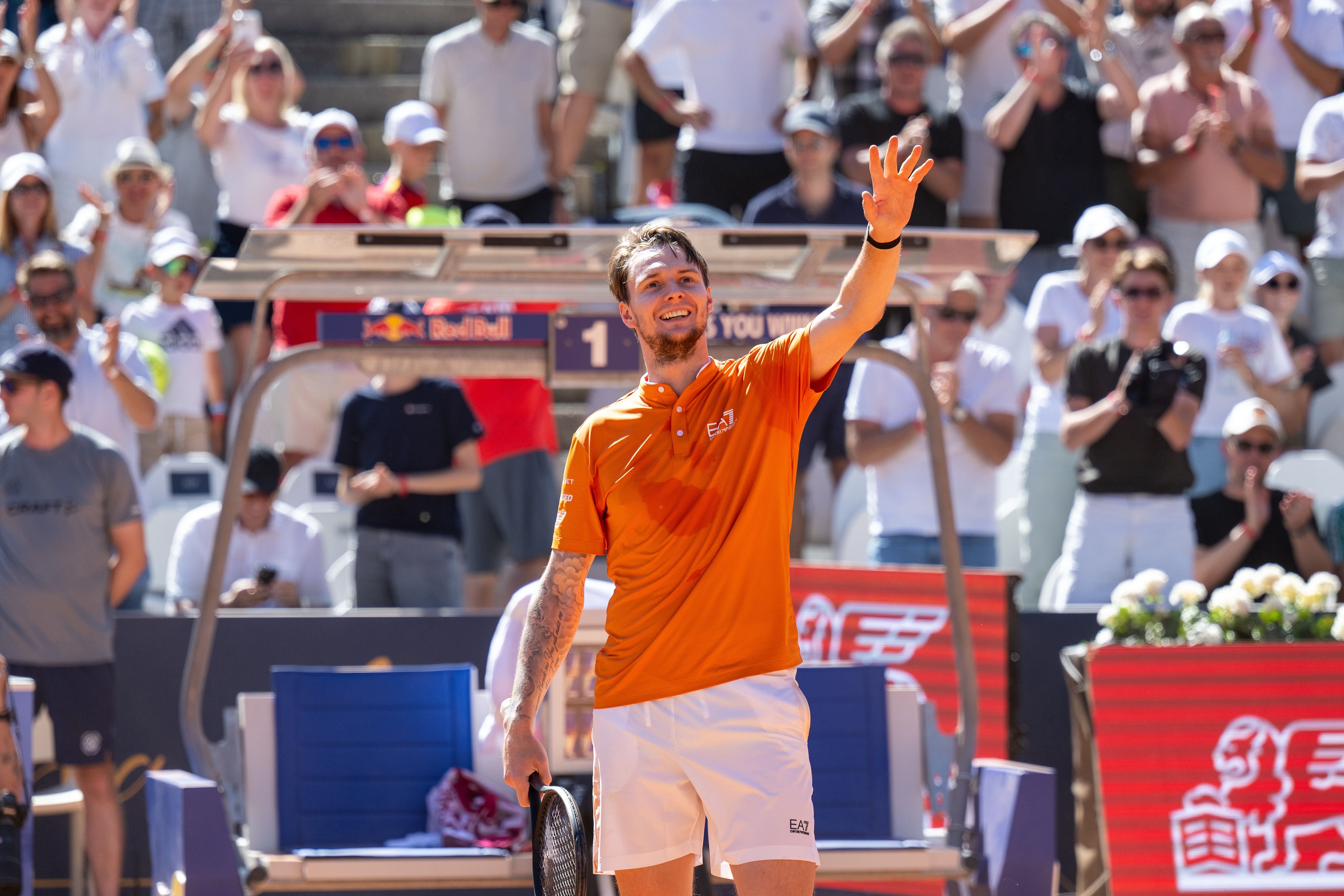 Ein Tennisspieler im orangenen Shirt jubelt auf dem Platz vor applaudierendem Publikum.