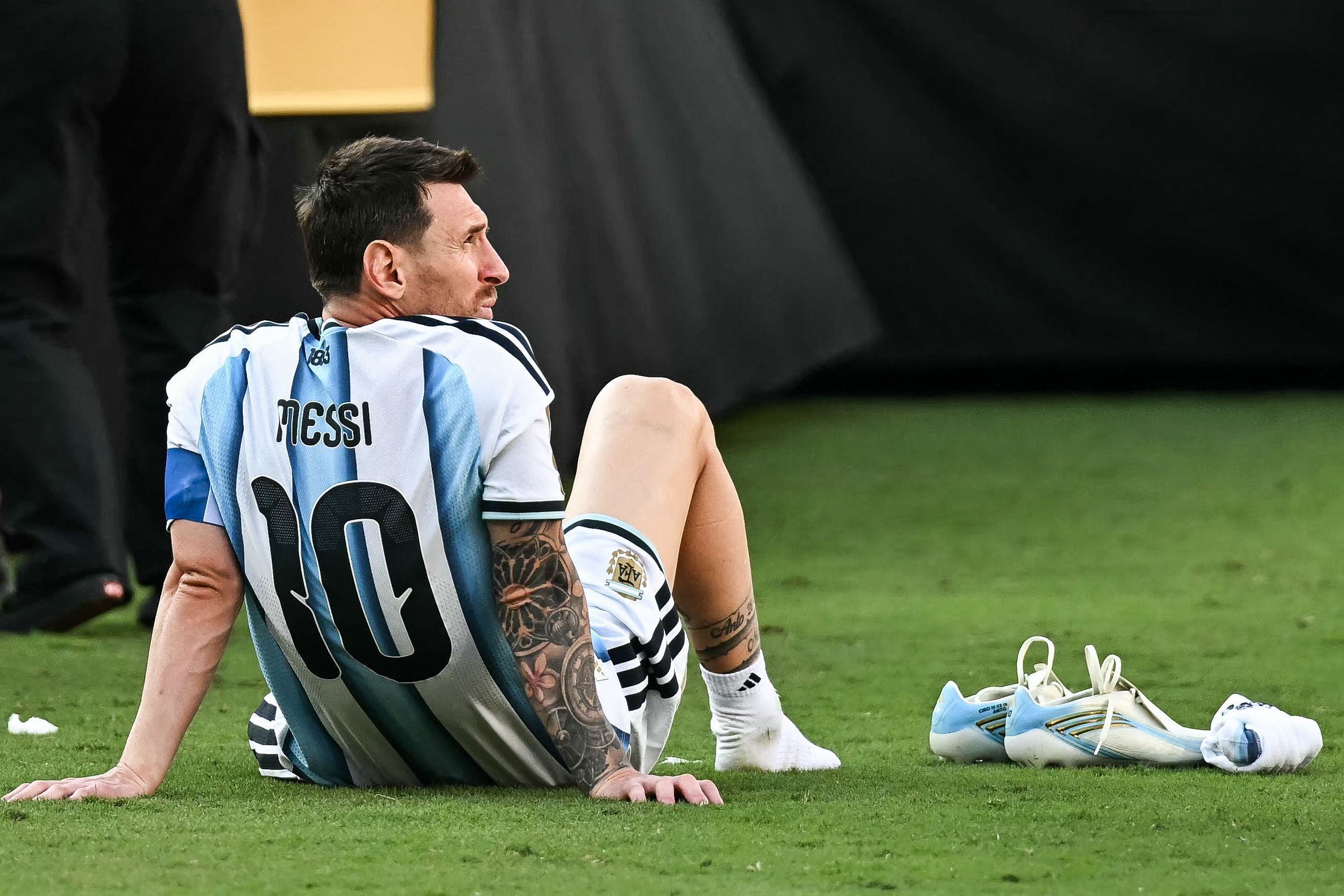 Ein argentinischer Fußballspieler sitzt traurig barfuß auf dem Rasen, seine Schuhe liegen daneben.