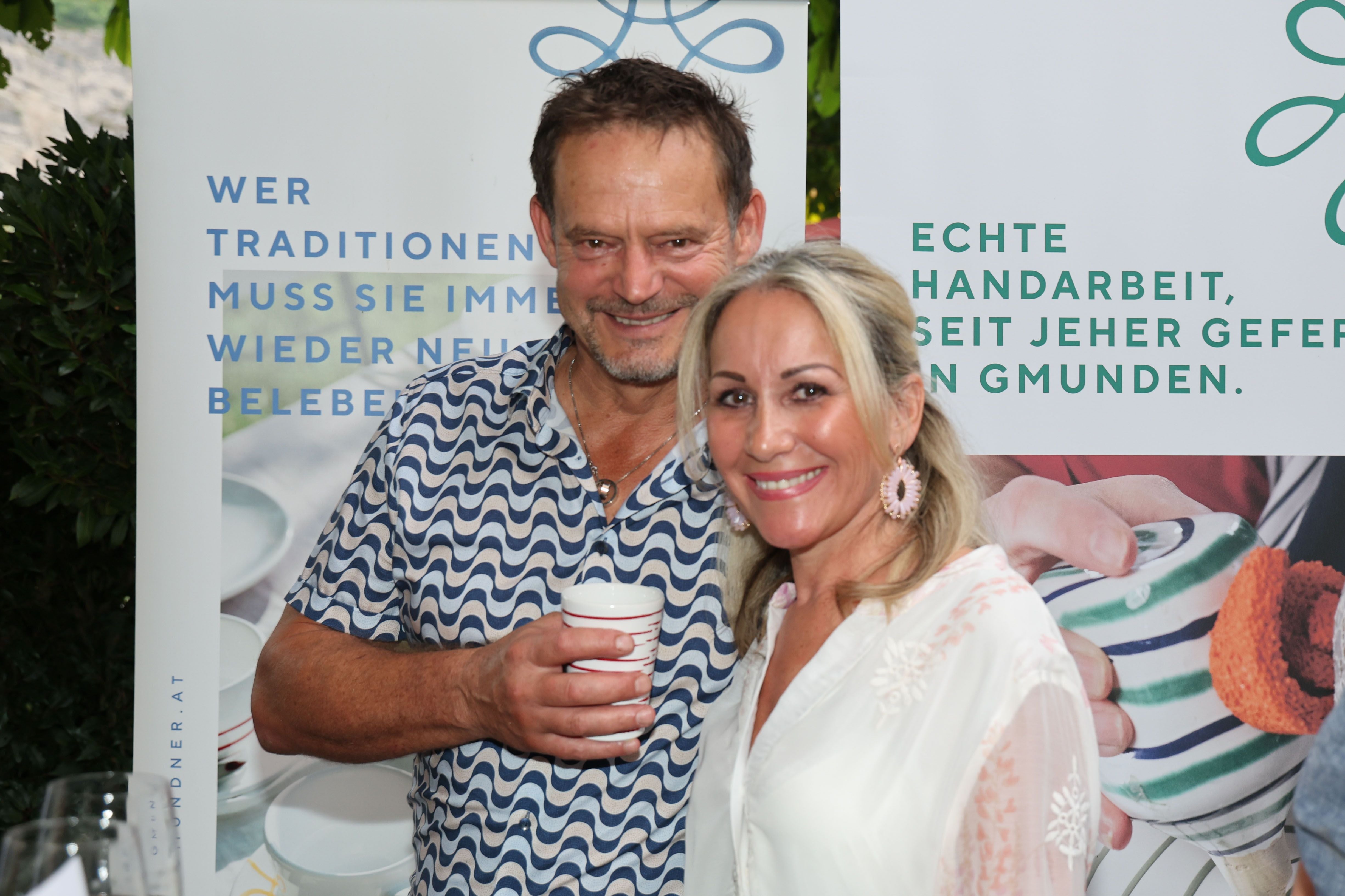 Ein Mann und eine Frau lächeln gemeinsam in die Kamera; der Mann hält einen Becher.