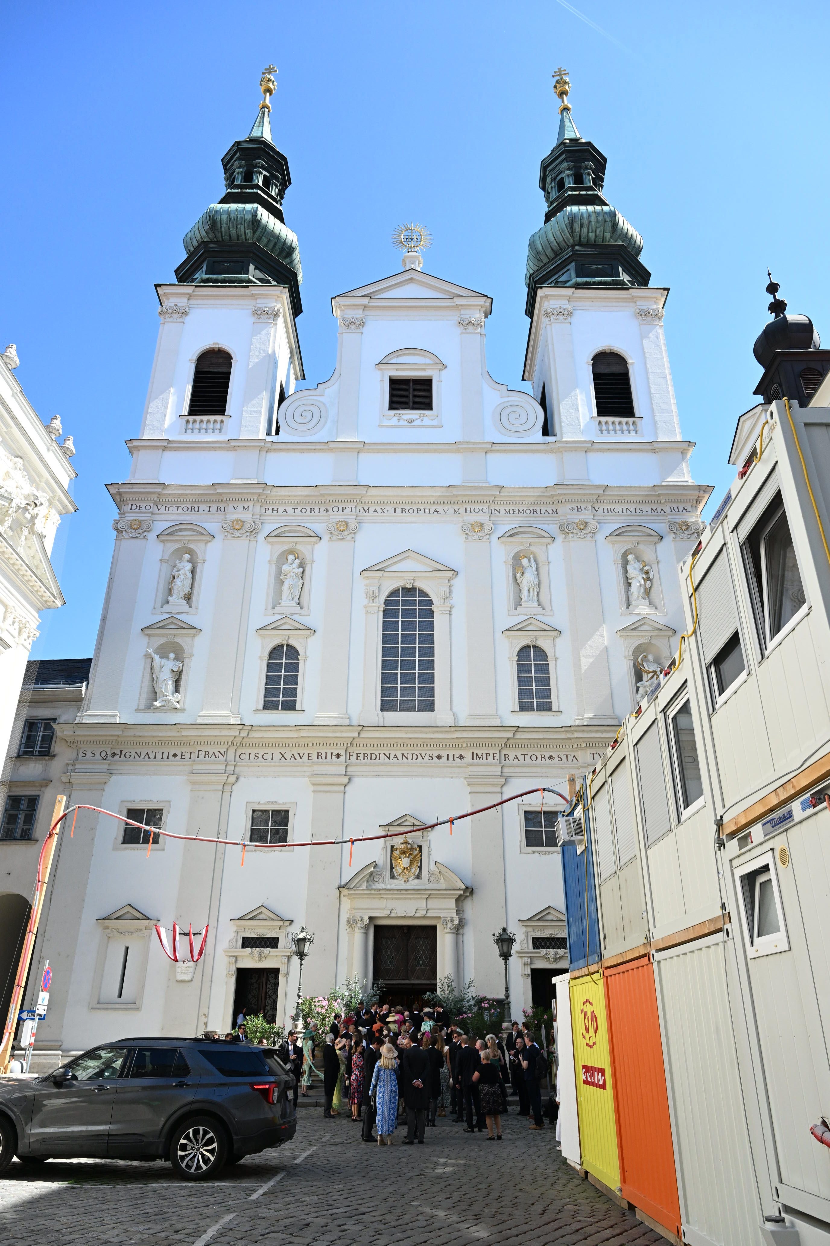 Menschen in festlicher Kleidung vor einer barocken Kirche bei sonnigem Wetter.