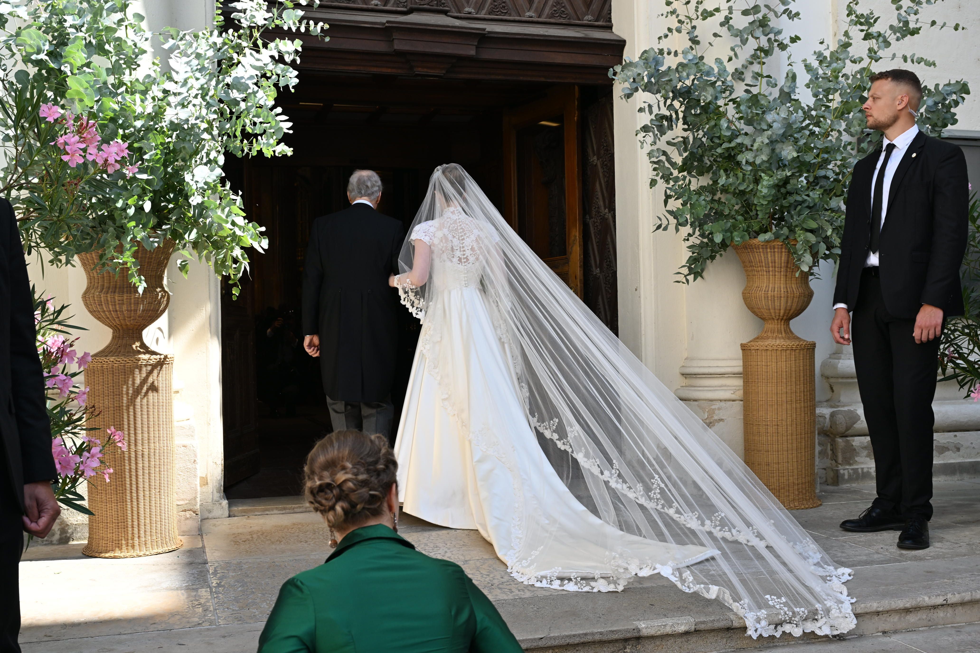 Braut im weißen Kleid mit langem Schleier betritt mit Begleitung eine Kirche bei einer Hochzeit.