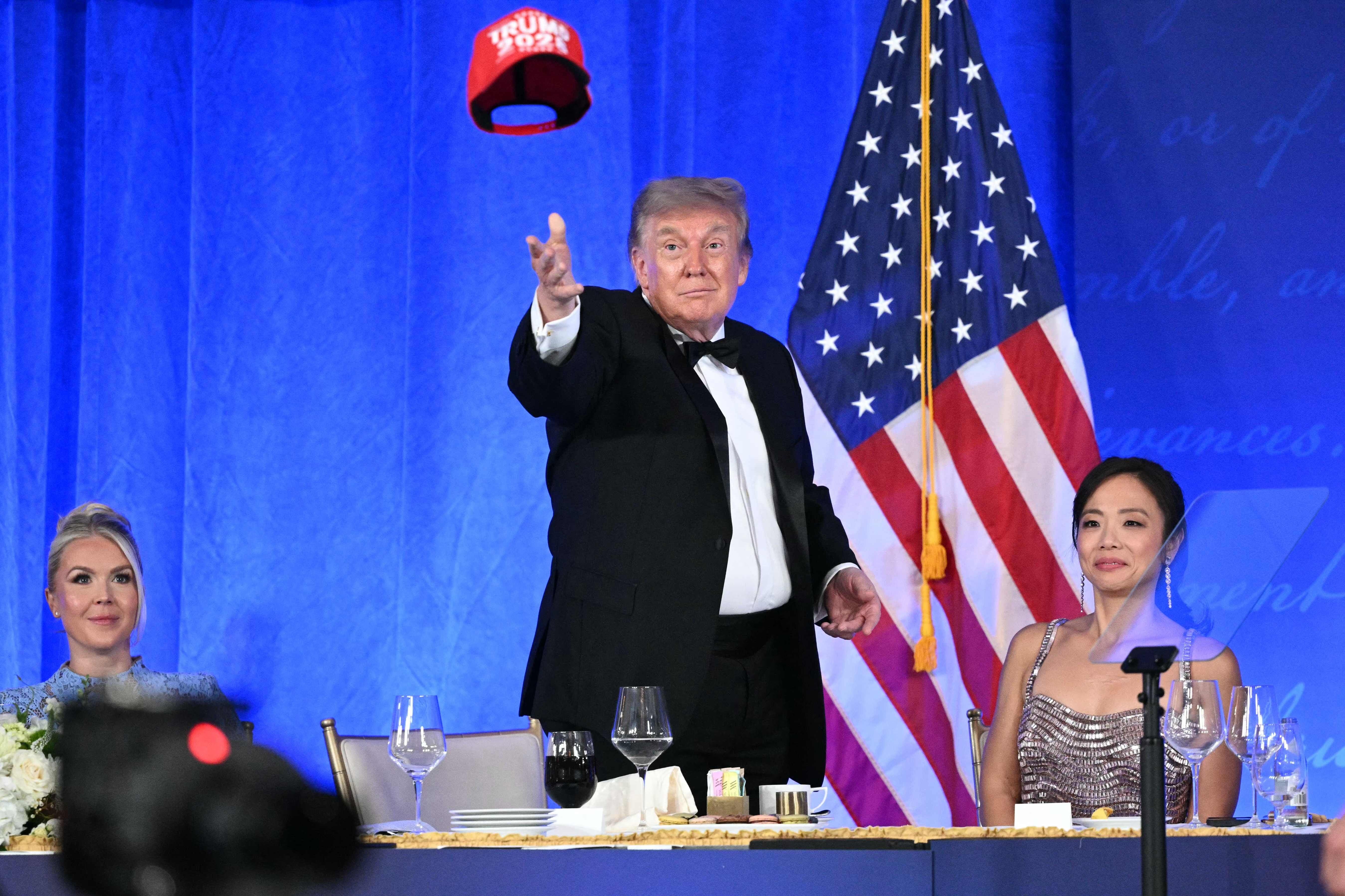 Ein Mann im Smoking wirft eine rote Kappe mit "Trump 2028" vor einer US-Flagge in einem Saal.