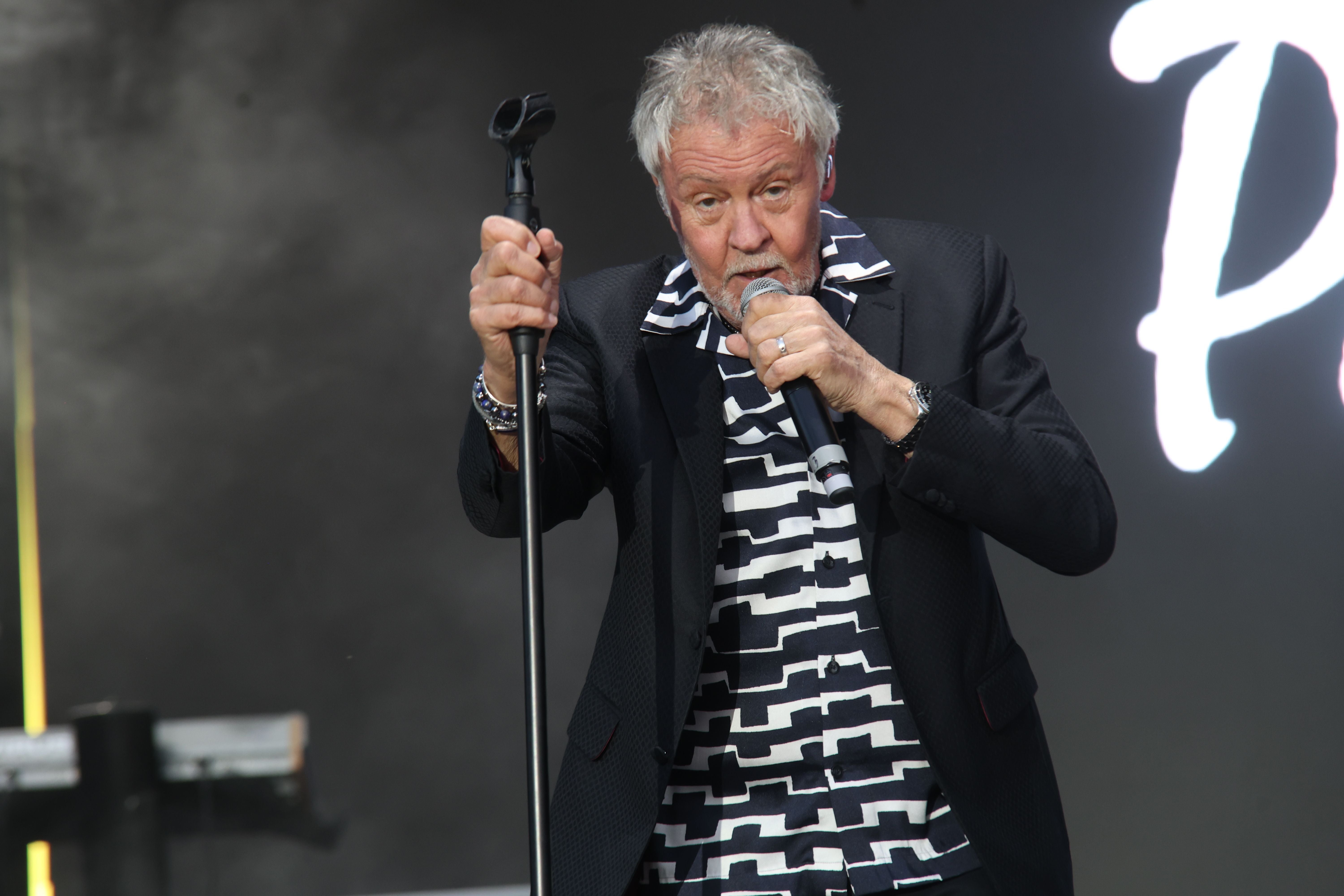 Ein älterer Mann singt auf der Bühne in ein Mikrofon und hält einen Mikrofonständer fest.