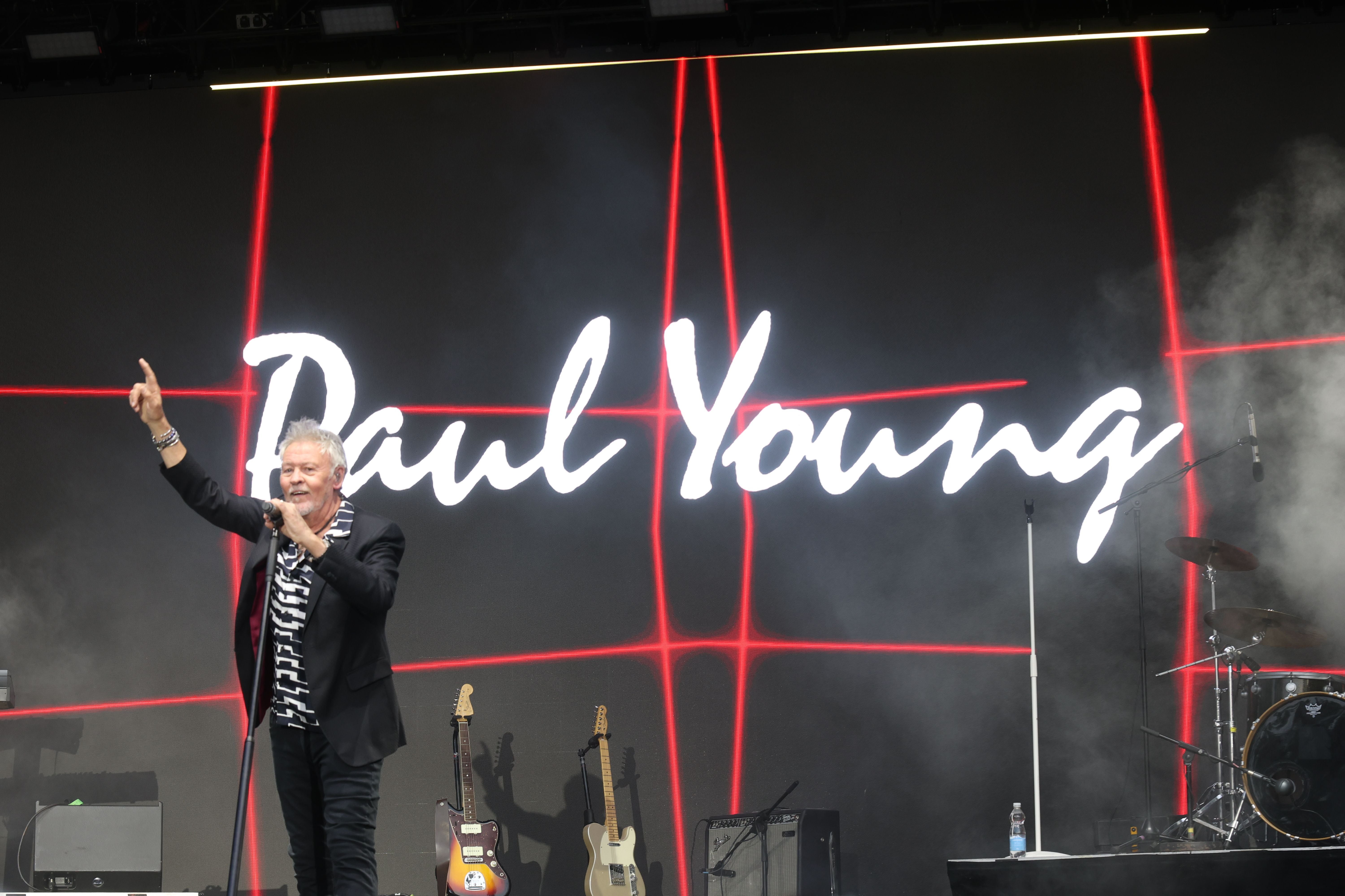 Sänger auf Bühne vor großem Bildschirm mit der Aufschrift "Paul Young", Instrumente und Nebel.